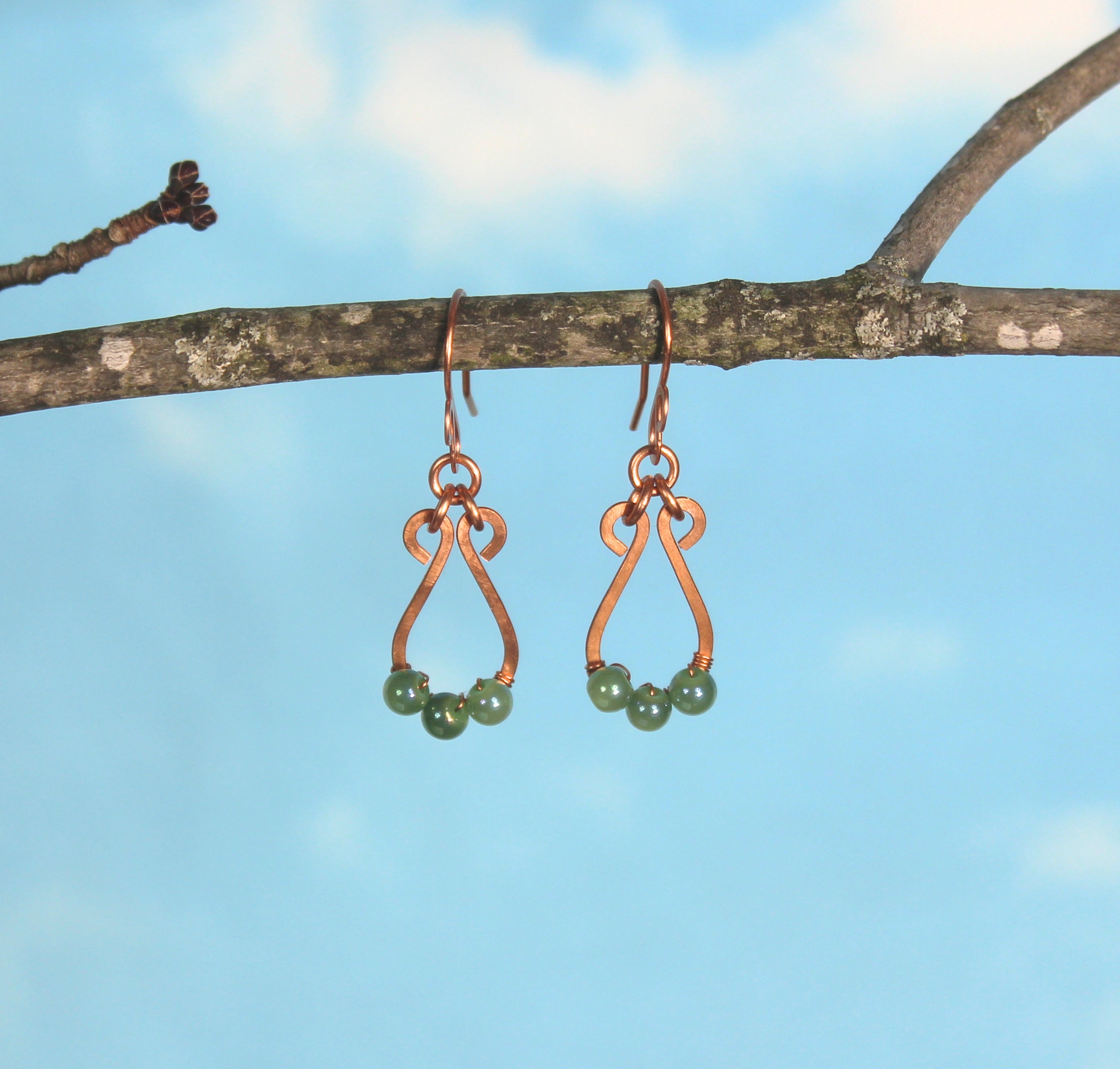 Hammered copper teardrop earrings with small green bead accents hanging on a branch against a blue sky.