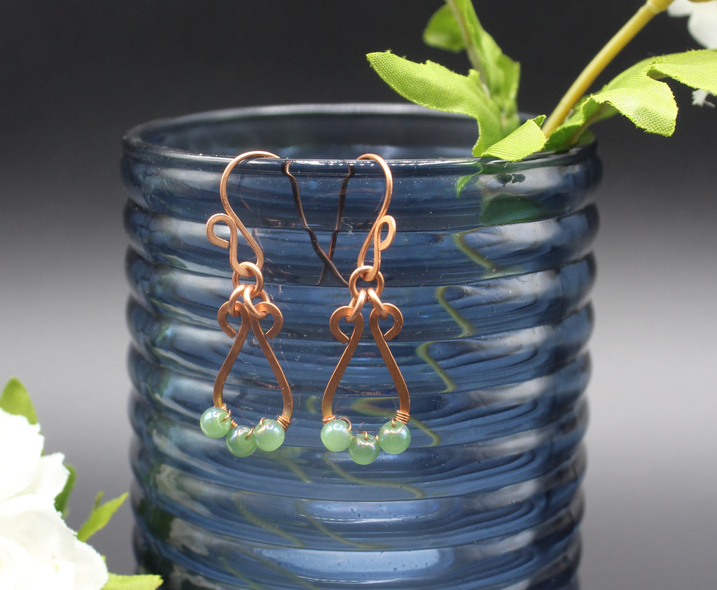 Hammered copper teardrop earrings with small green bead accents in front of a blue glass vase.