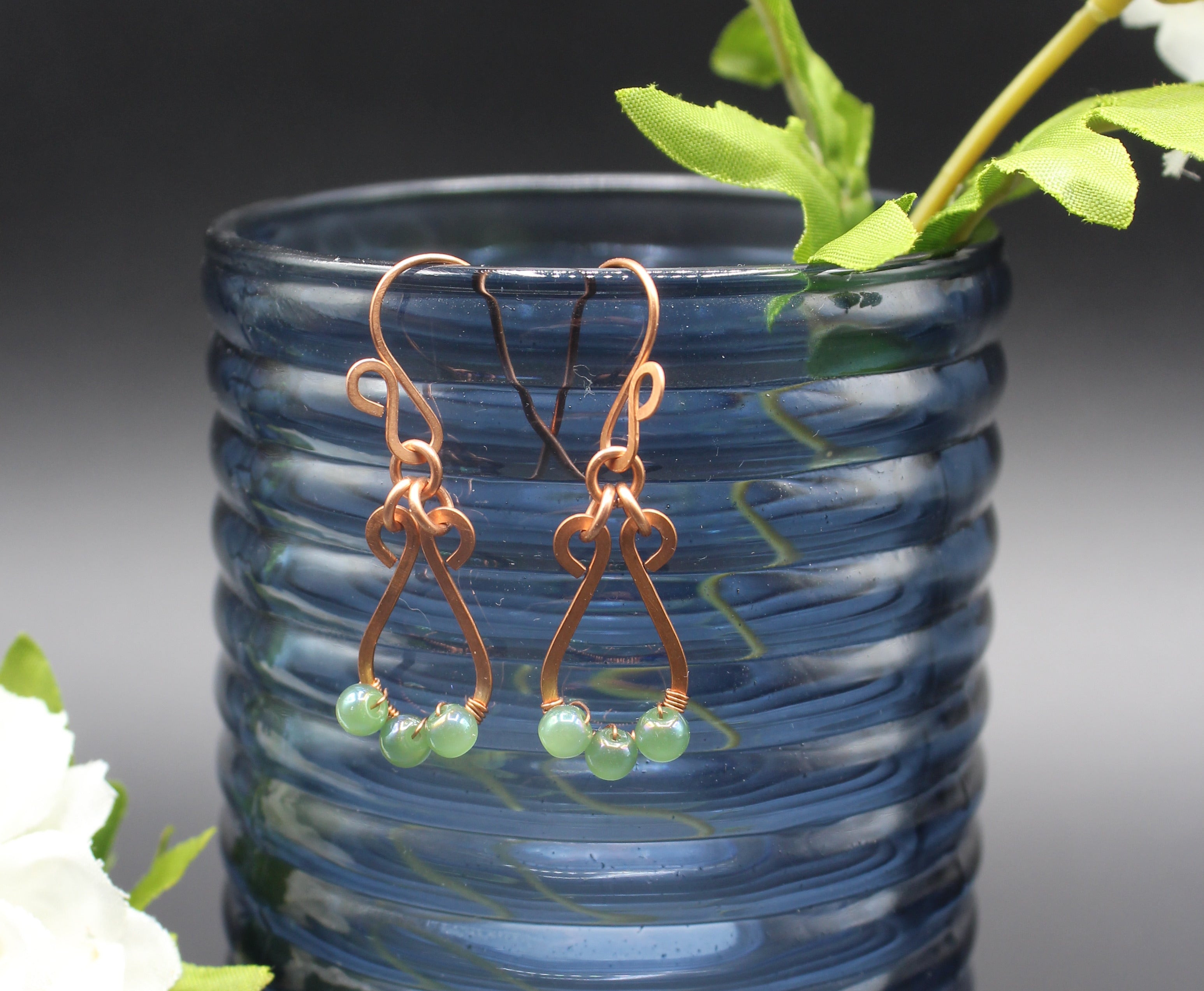 Hammered copper teardrop earrings with small green bead accents in front of a blue glass vase.