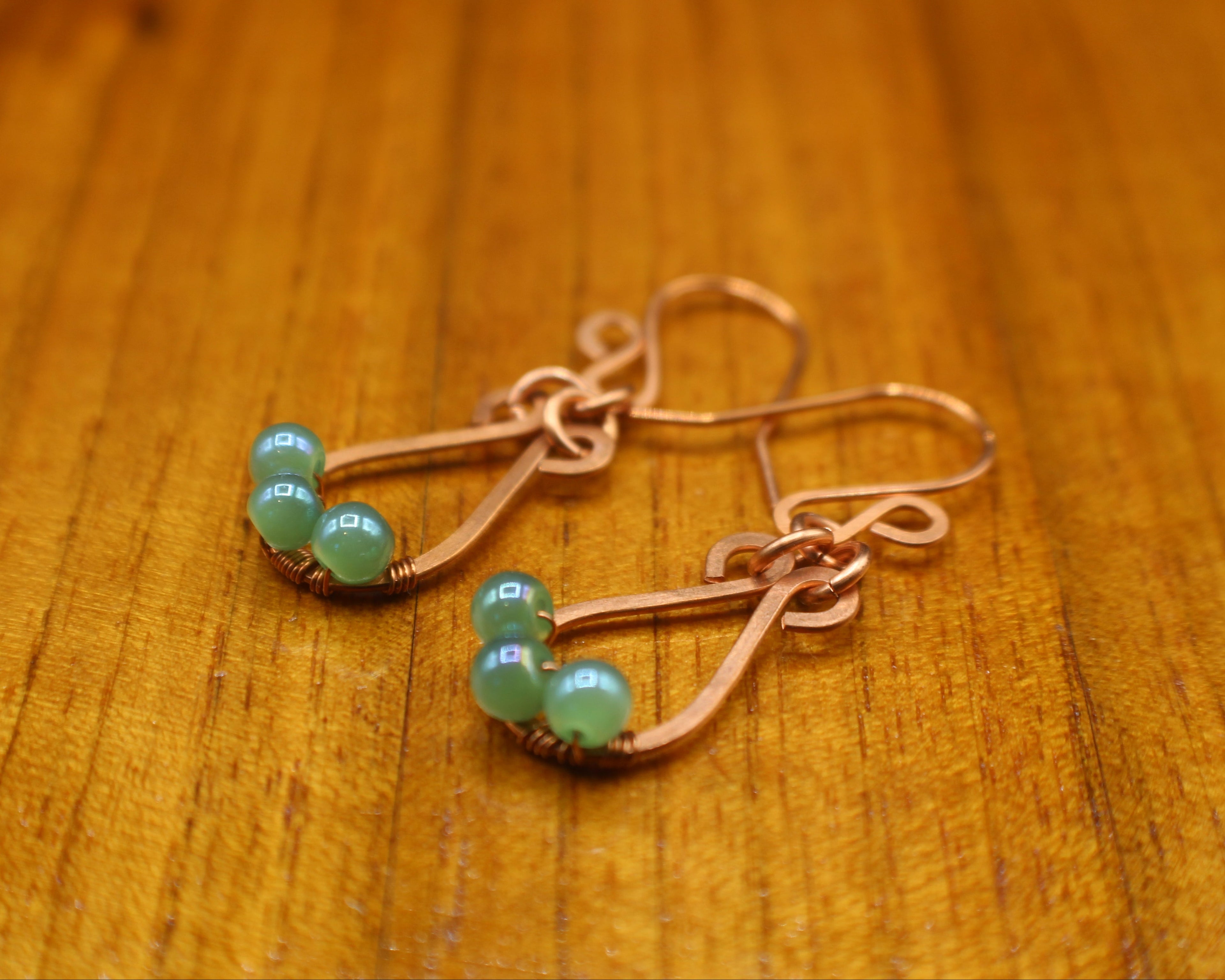 Hammered copper teardrop earrings with small green bead accents on a wooden surface