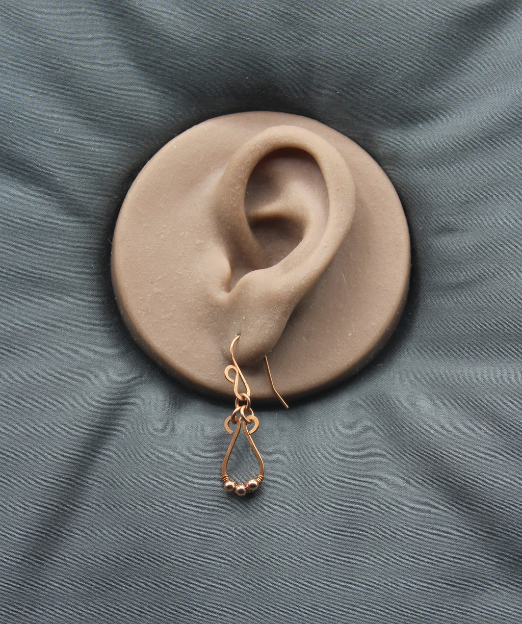 Hammered copper teardrop earring with tiny wrapped beads on a clay model of an ear with a gray background
