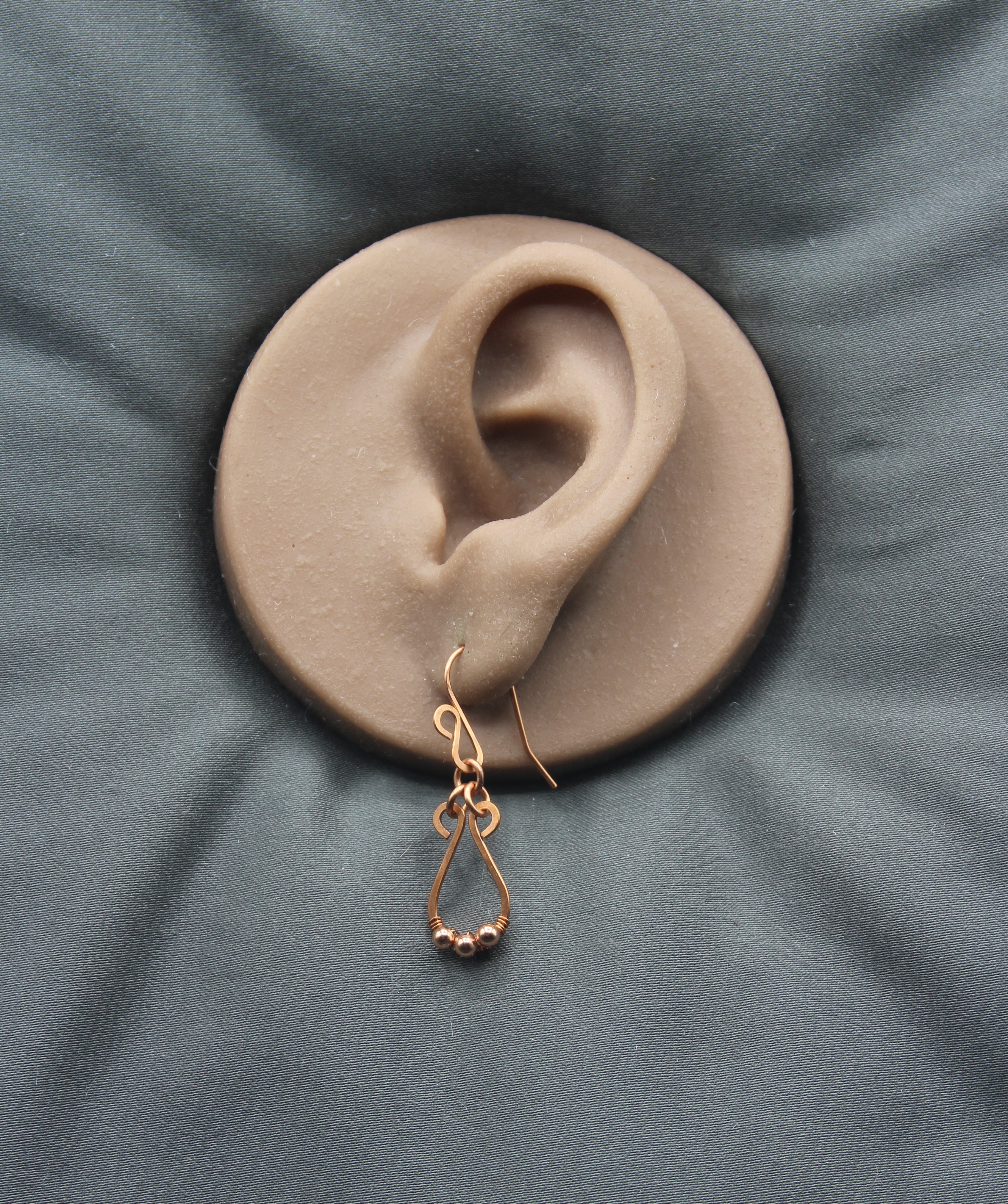 Hammered copper teardrop earring with tiny wrapped beads on a clay model of an ear with a gray background