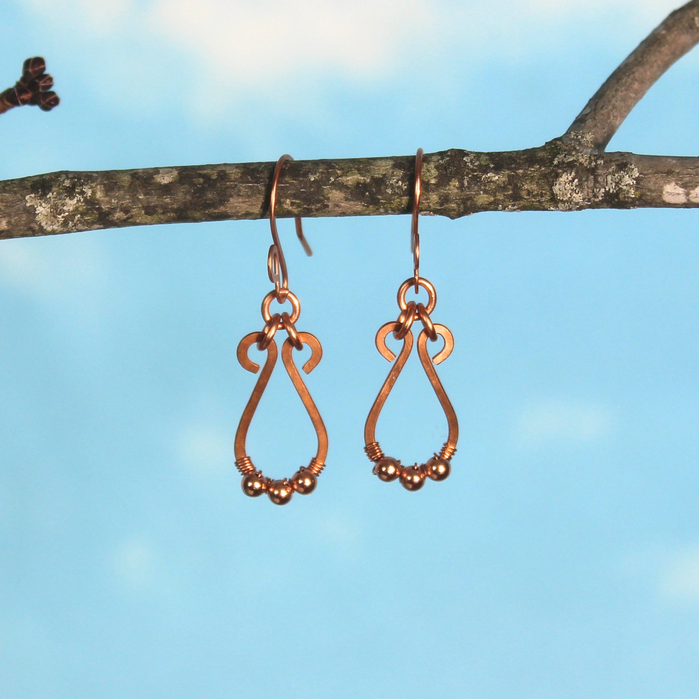 Hammered copper teardrop earrings with tiny wrapped beads hanging from a branch with a blue sky background