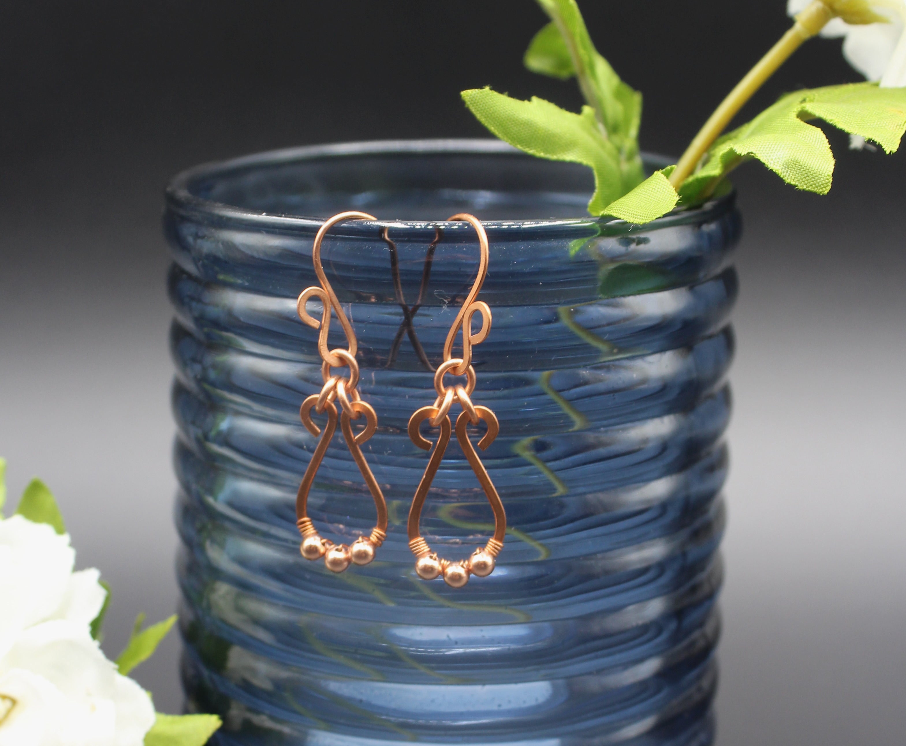 Hammered copper teardrop earrings with tiny wrapped beads on a gray background on a blue glass vase with greenery