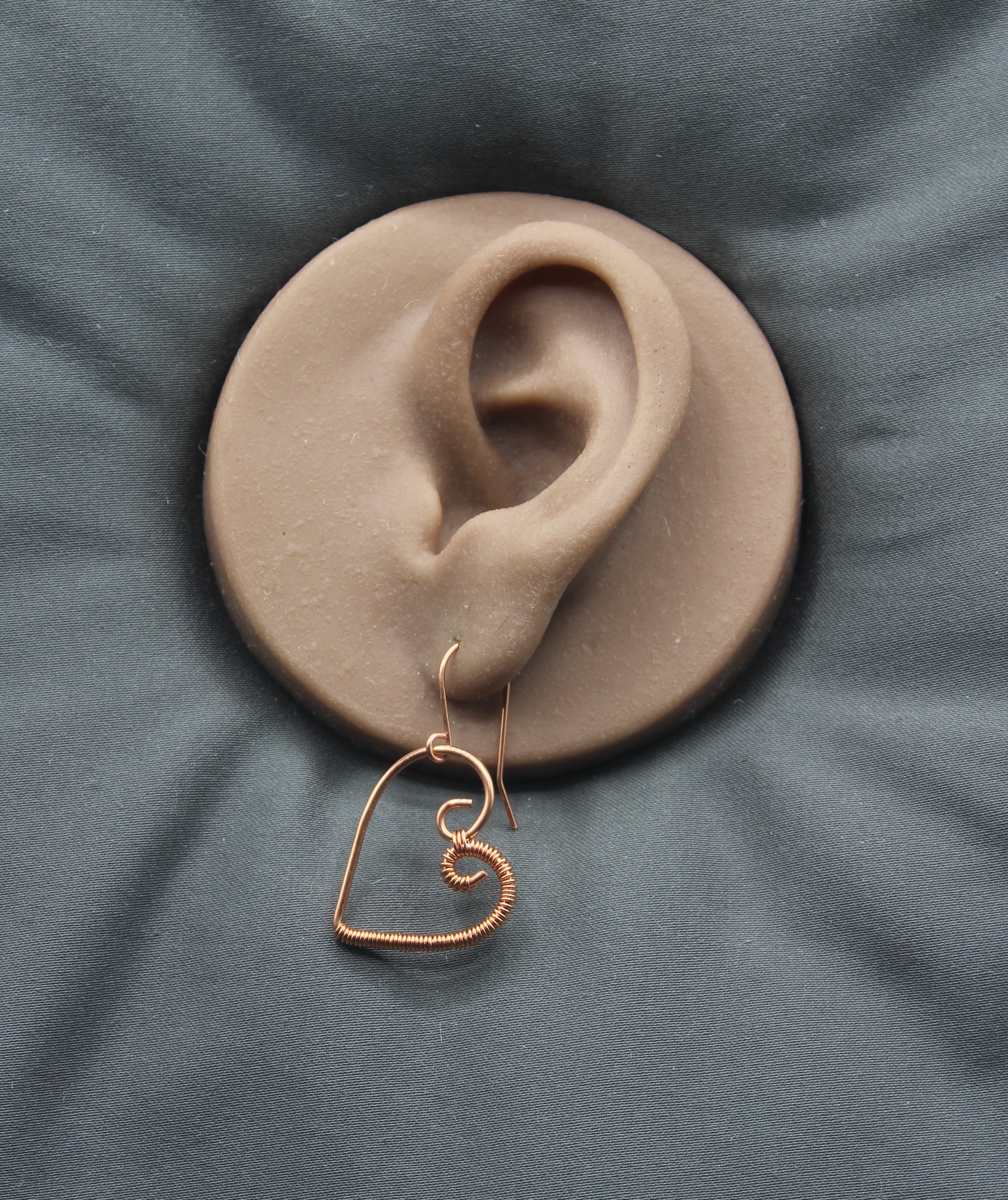 Heart-shaped copper wire earring on a clay model of an ear against a gray background