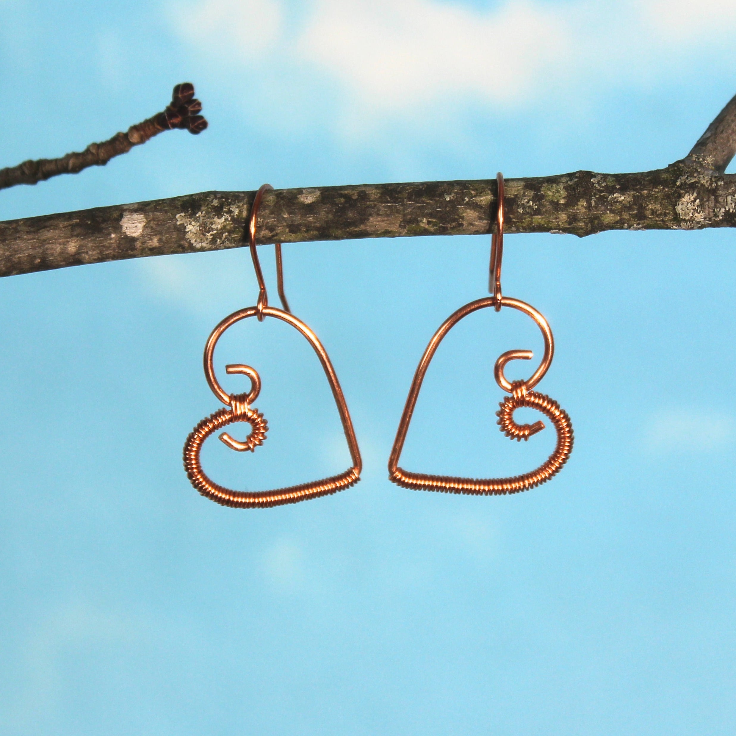 Copper wire heart-shaped earrings hanging from a branch against a blue sky.