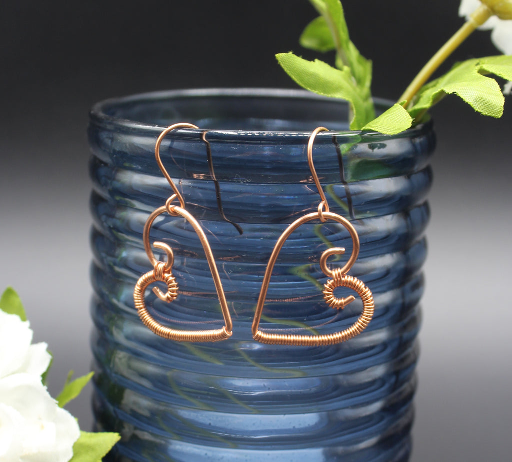 Copper wire heart-shaped earrings hanging on a blue glass vase with greenery.