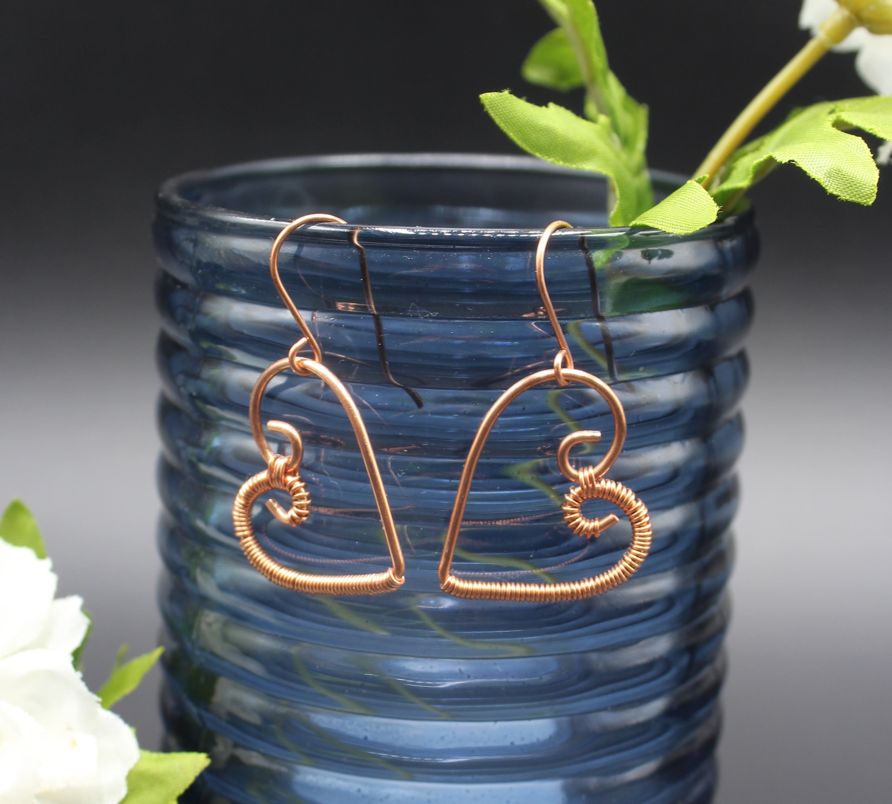 Copper wire heart-shaped earrings hanging on a blue glass vase with greenery.