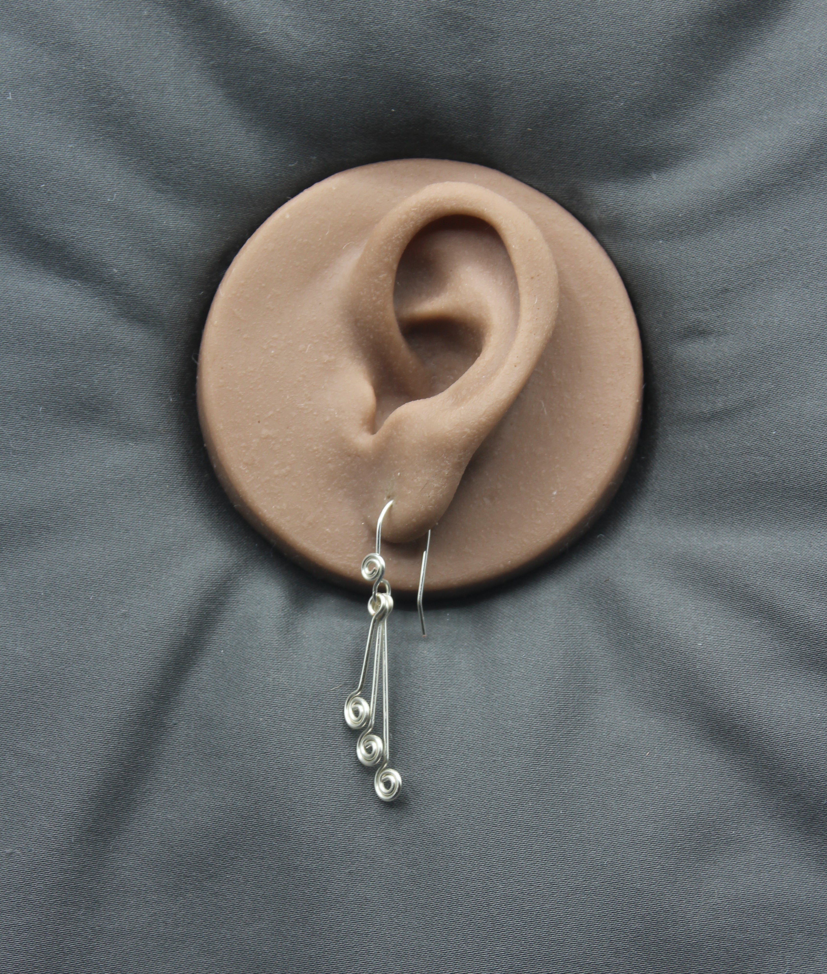 Silver wire earrings  with hanging triple spirals displayed on a clay model of an ear against a gray background