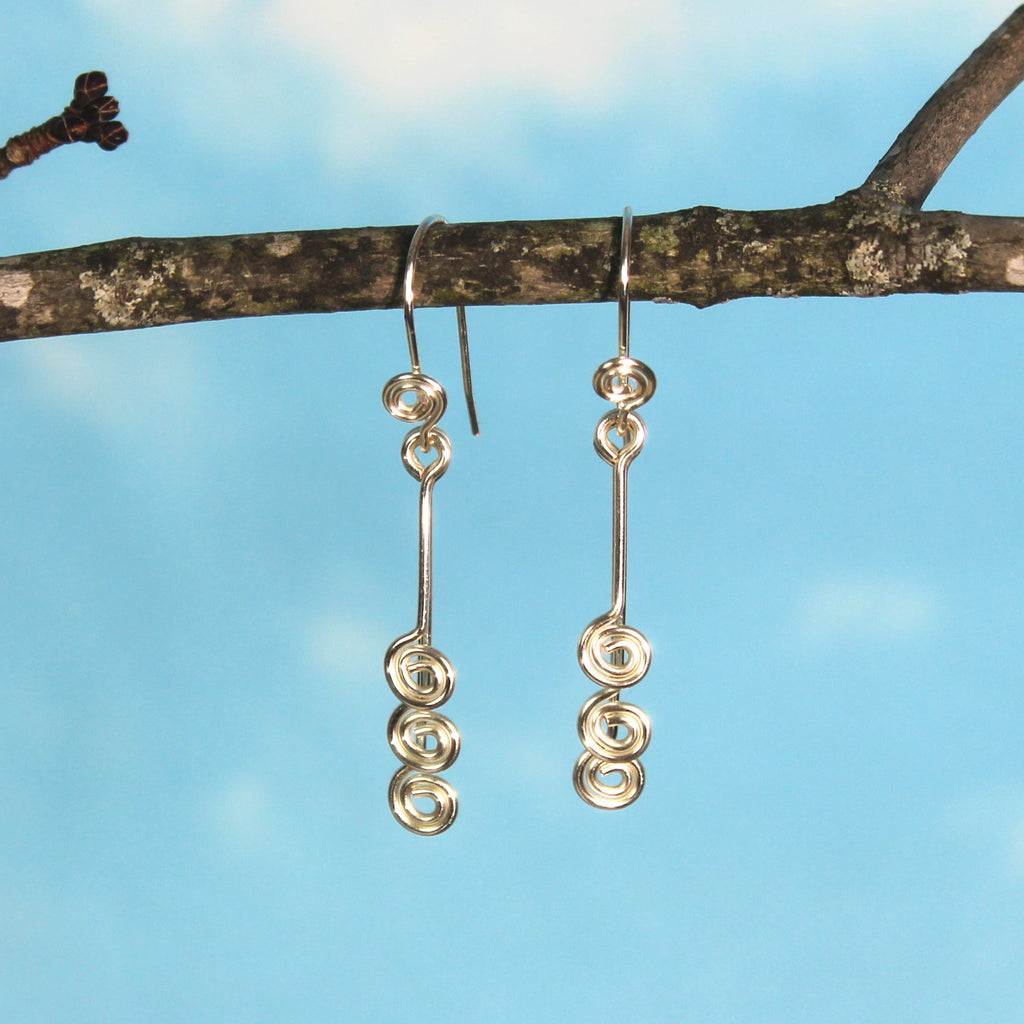 Pair of silver wire earrings with small spirals hanging from a branch with a blue sky background