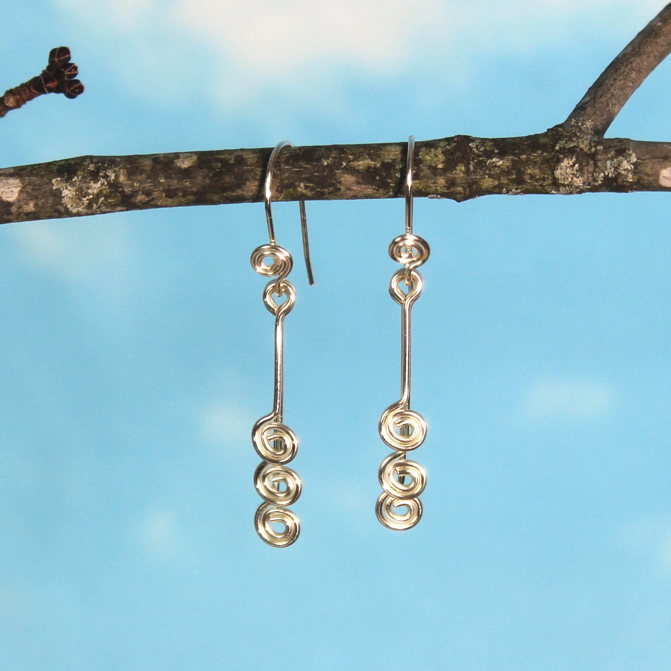 Pair of silver wire earrings with small spirals hanging from a branch with a blue sky background