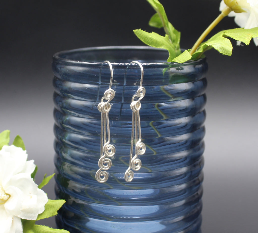 Silver wire earrings with small spirals hanging from a blue glass vase with flowers.