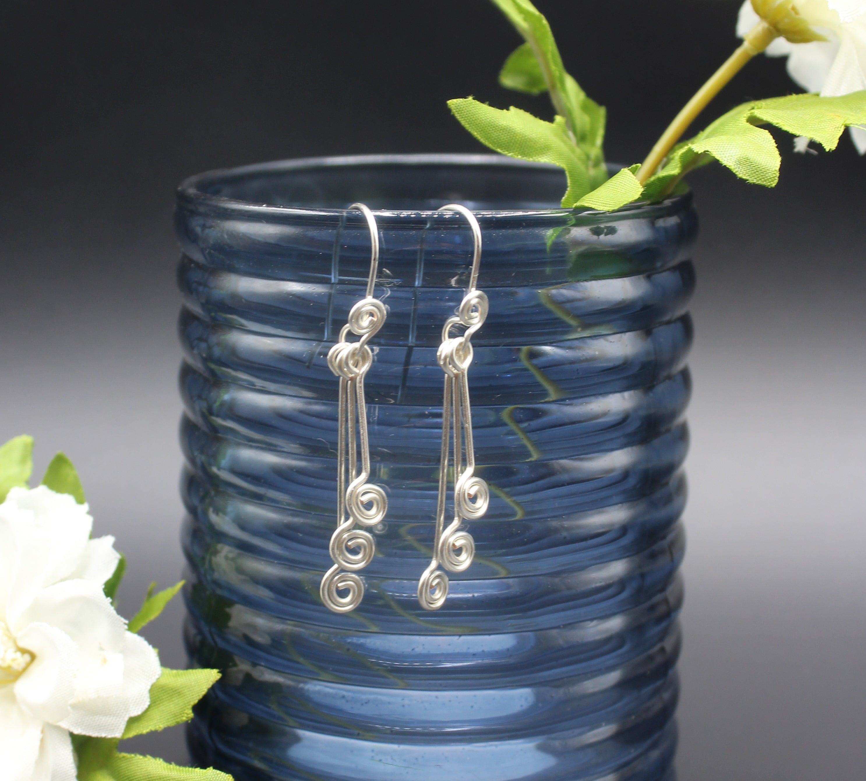 Silver wire earrings with small spirals hanging from a blue glass vase with flowers.