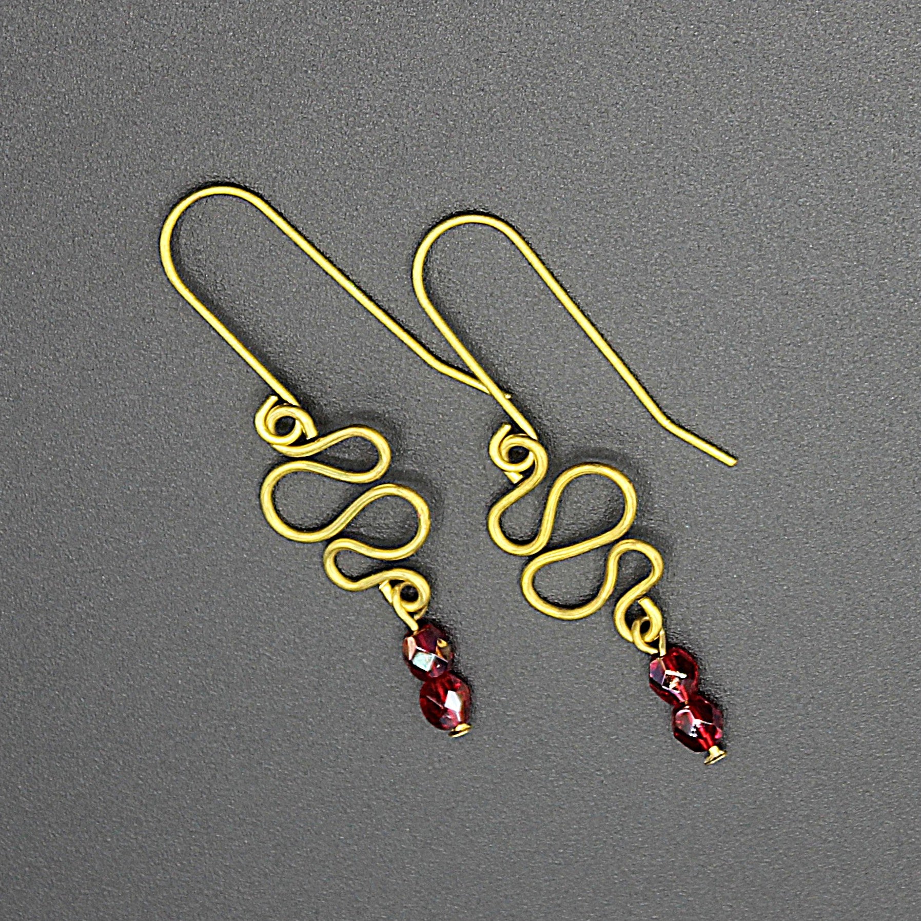 Freeform brass wire earrings with red faceted beads on a gray background