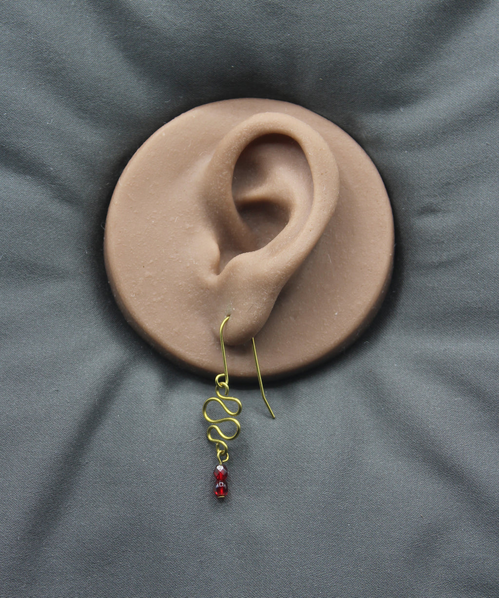 Freeform brass wire earring with red faceted beads on a clay ear model against a gray background