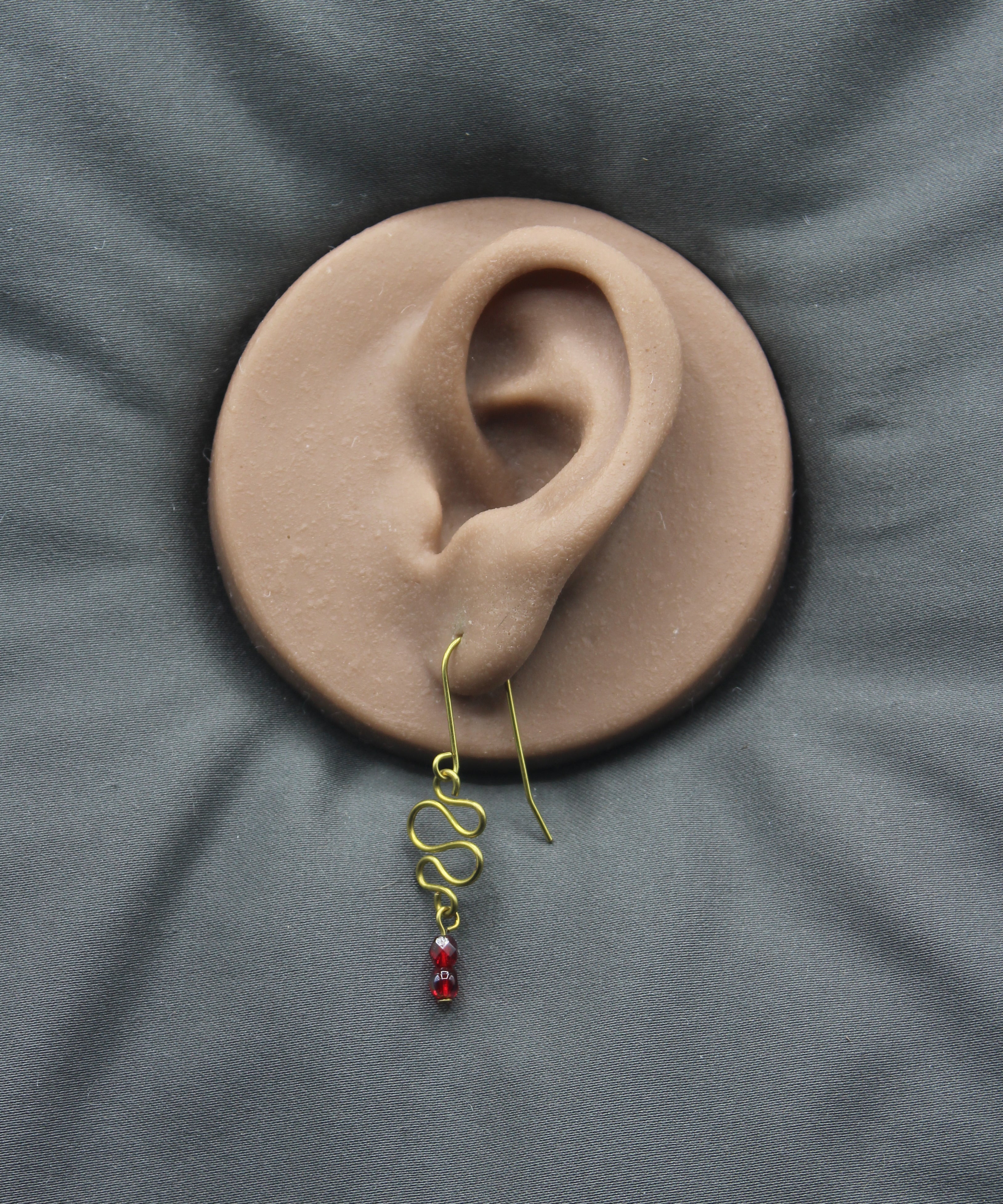 Freeform brass wire earring with red faceted beads on a clay ear model against a gray background