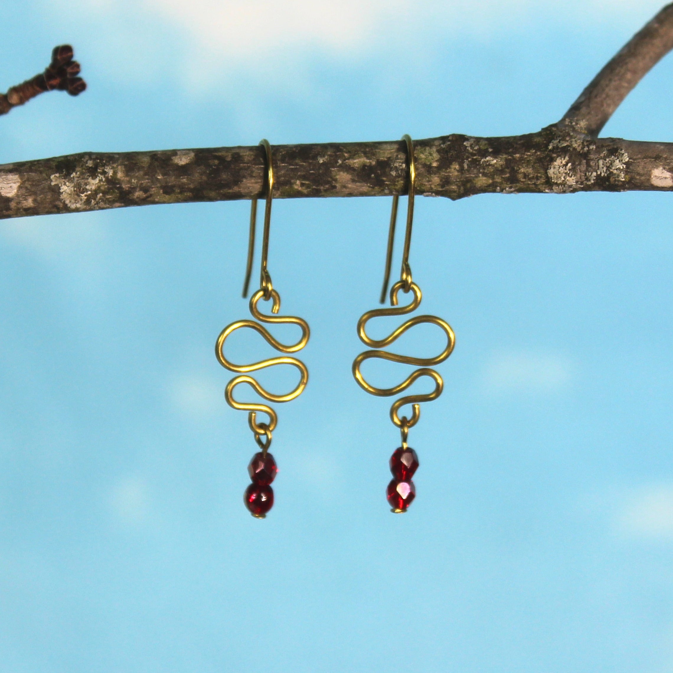 Freeform brass wire earrings with red faceted beads hanging from a branch against a blue sky.