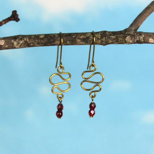 Freeform brass wire earrings with red faceted beads hanging from a branch against a blue sky.