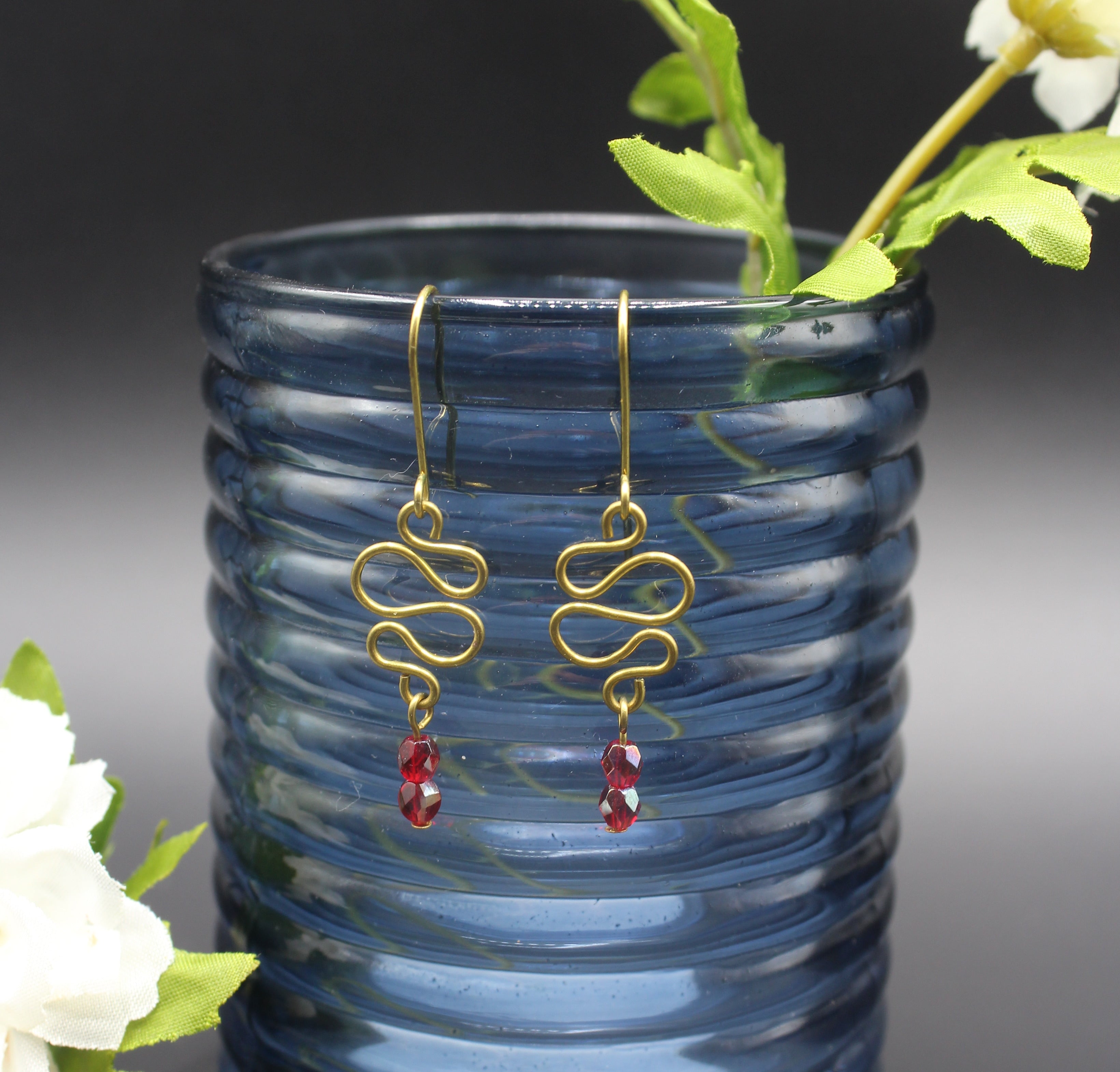 Freeform brass wire earrings with red faceted beads hanging from a blue glass vase.