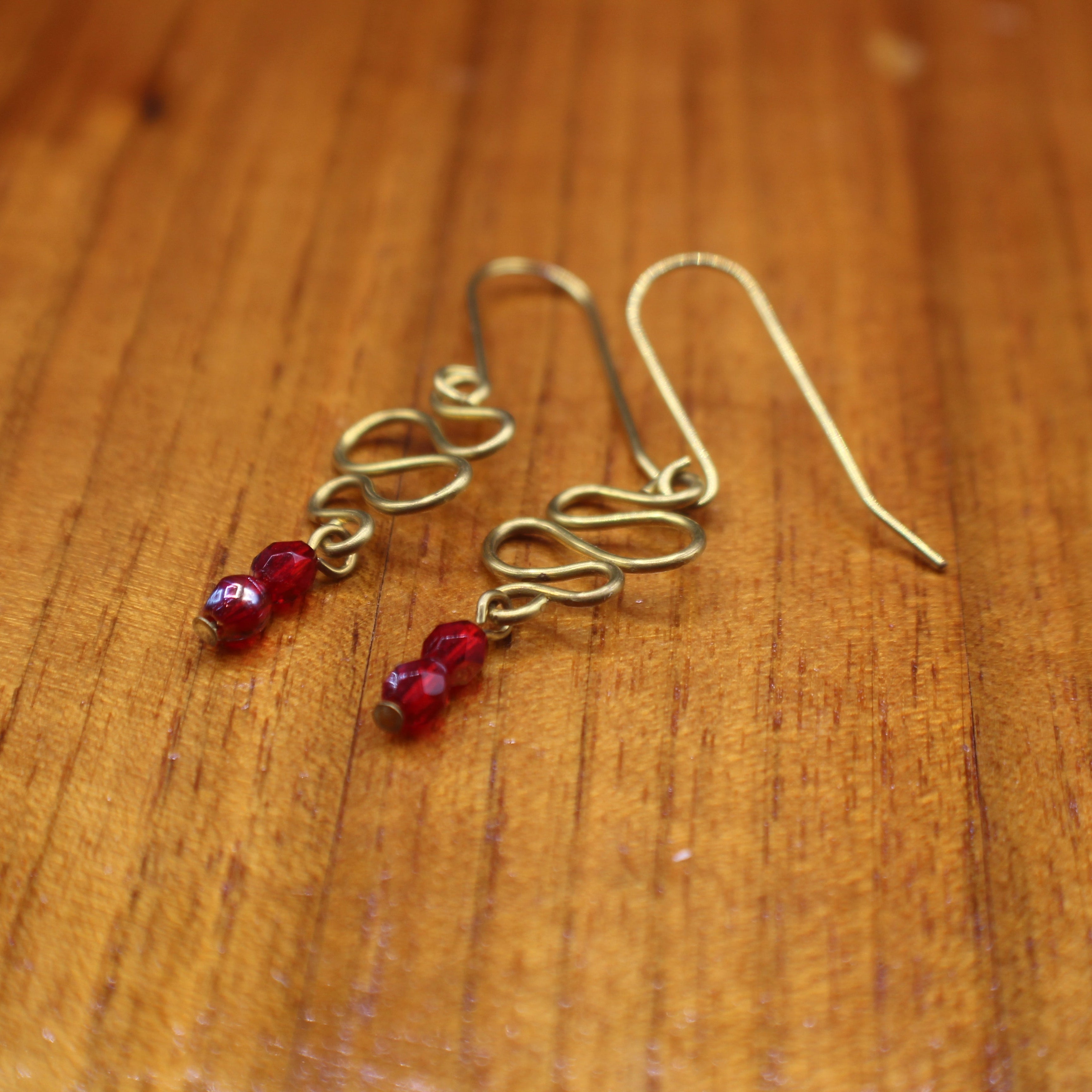 Freeform brass wire earrings with red faceted beads on a wooden surface