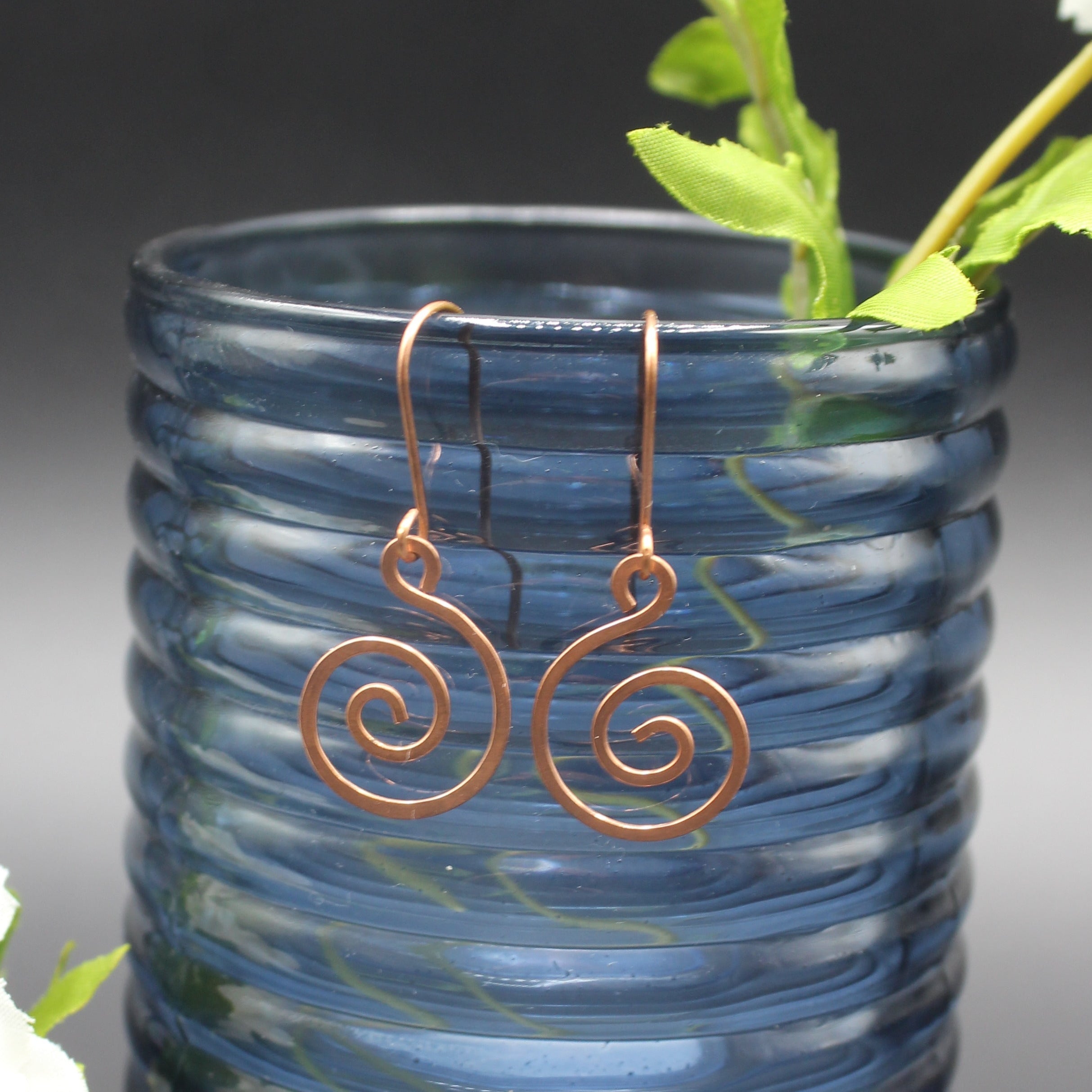 Copper spiral earrings hanging from a blue glass vase with greenery.