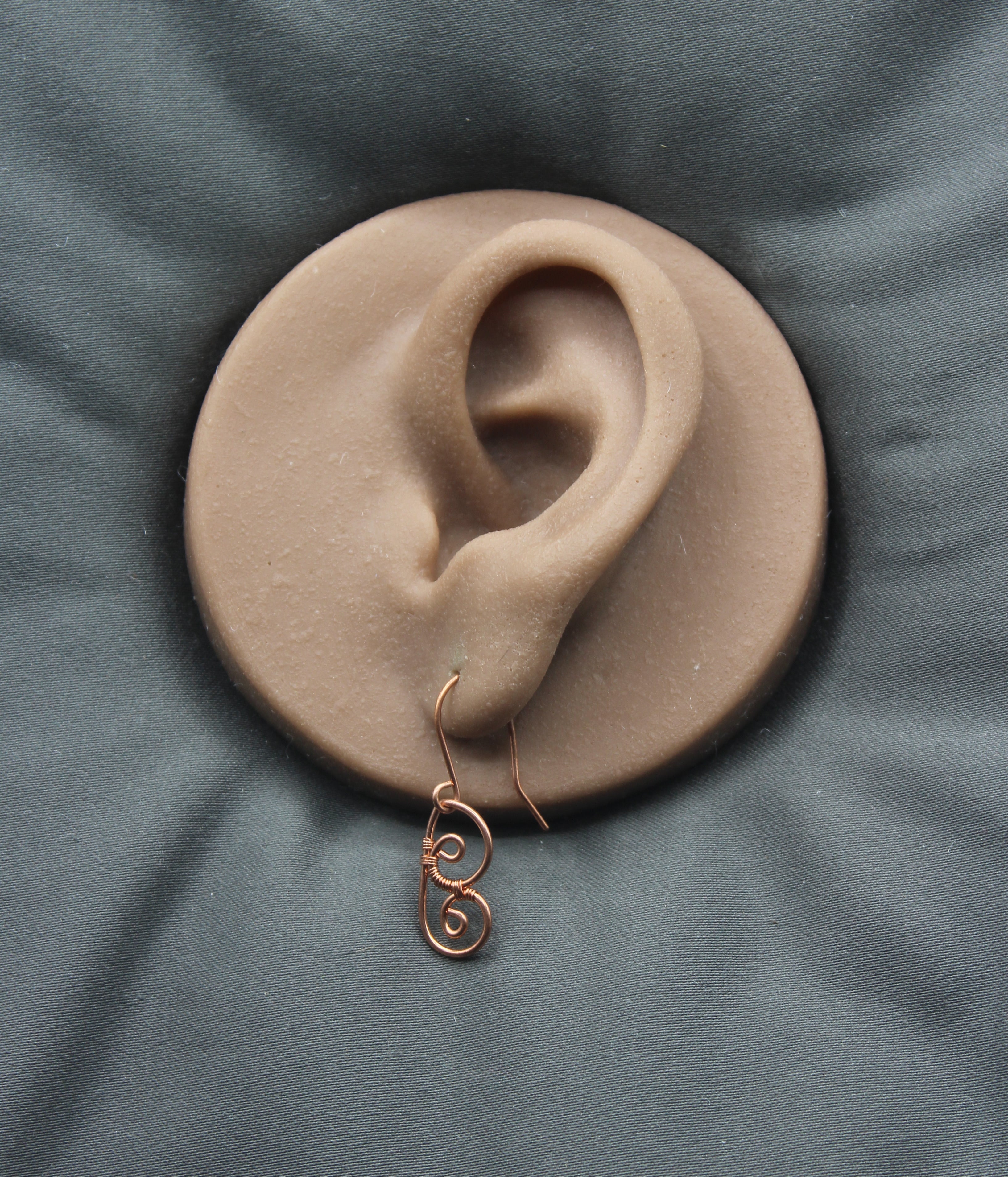 Copper wire earring shaped as an abstract butterfly wing, on a clay model of an ear against a gray background