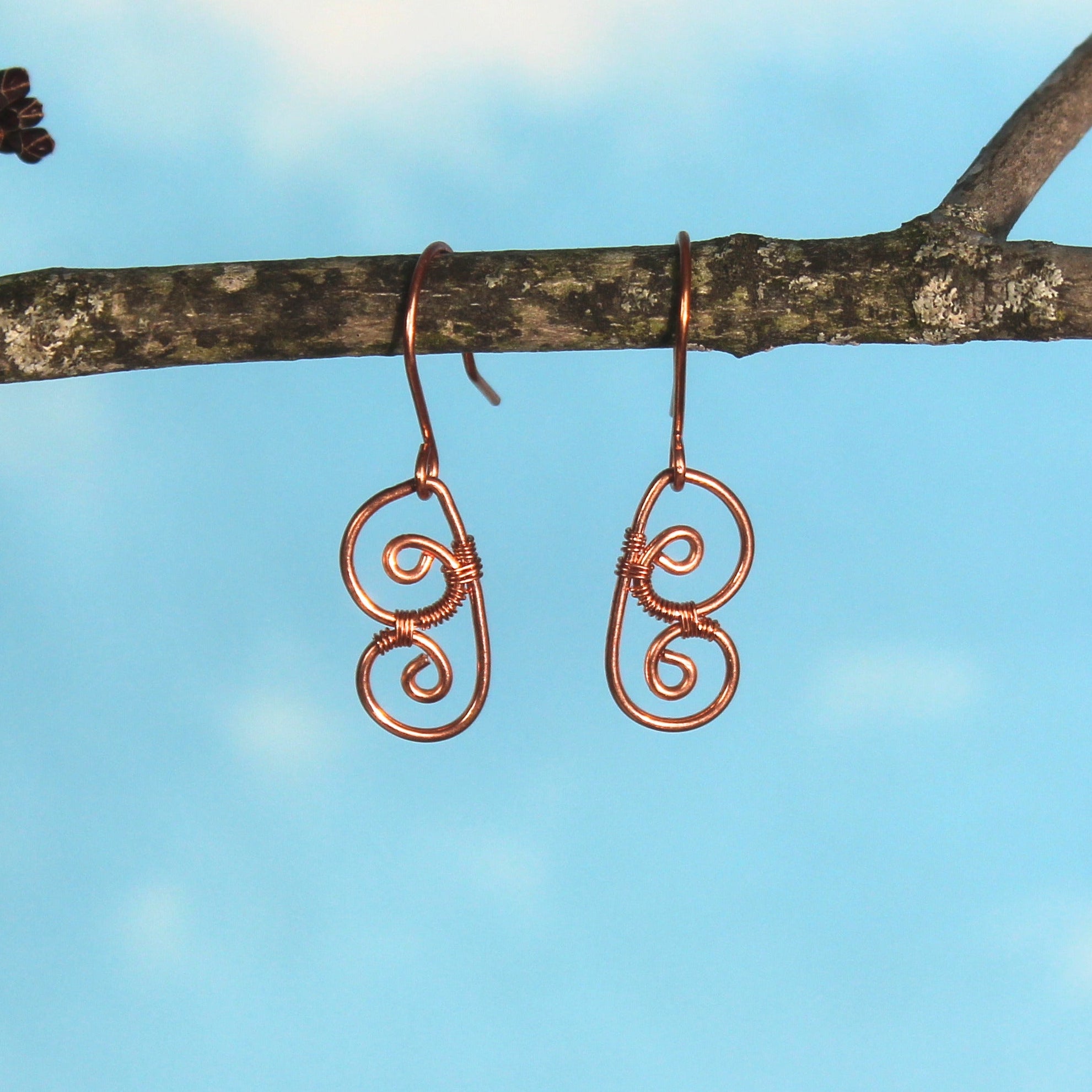 Copper wire earrings shaped as abstract butterfly wings, hanging from a branch against a blue sky.