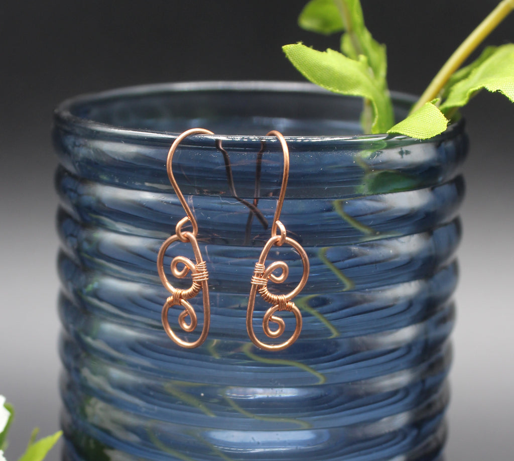 Copper wire earrings shaped as abstract butterfly wings hanging from a blue glass vase with green leaves.