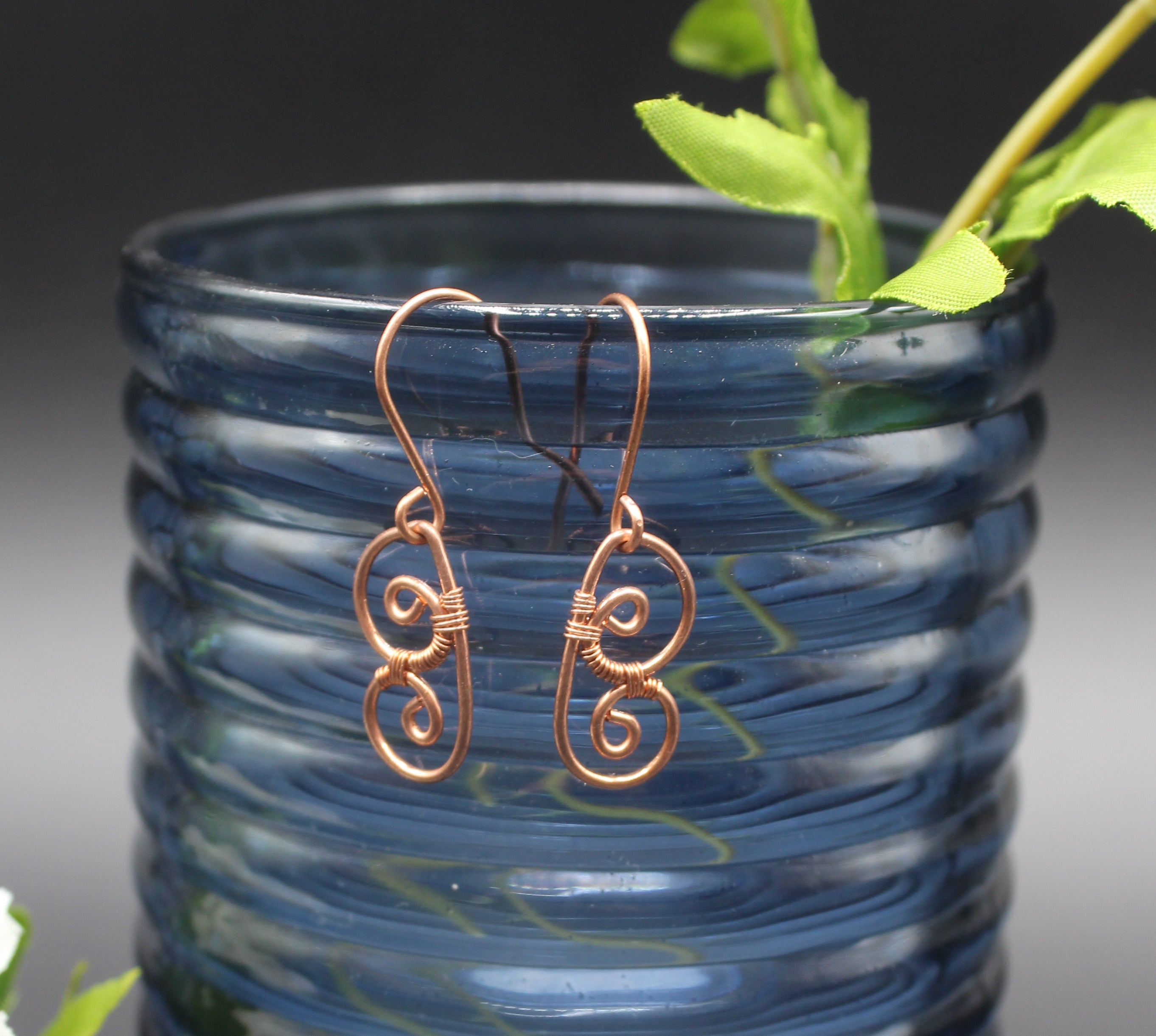 Copper wire earrings shaped as abstract butterfly wings hanging from a blue glass vase with green leaves.