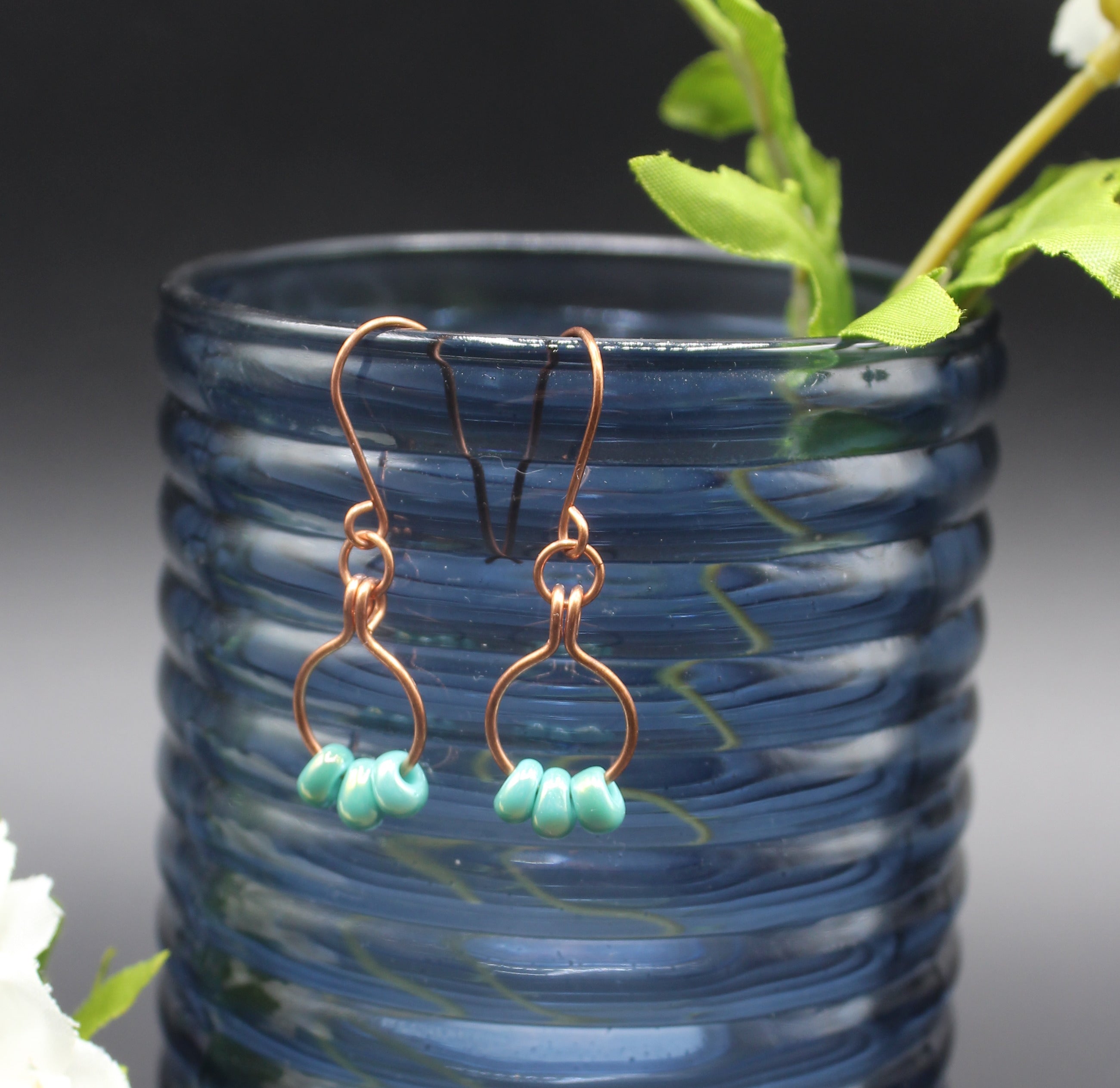 Copper wire earrings with green beads on small hoops hanging from a blue glass vase.