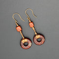 Pair of earrings with donut-shaped brownish-red stones that have striations, wrapped with copper  wire on a gray background