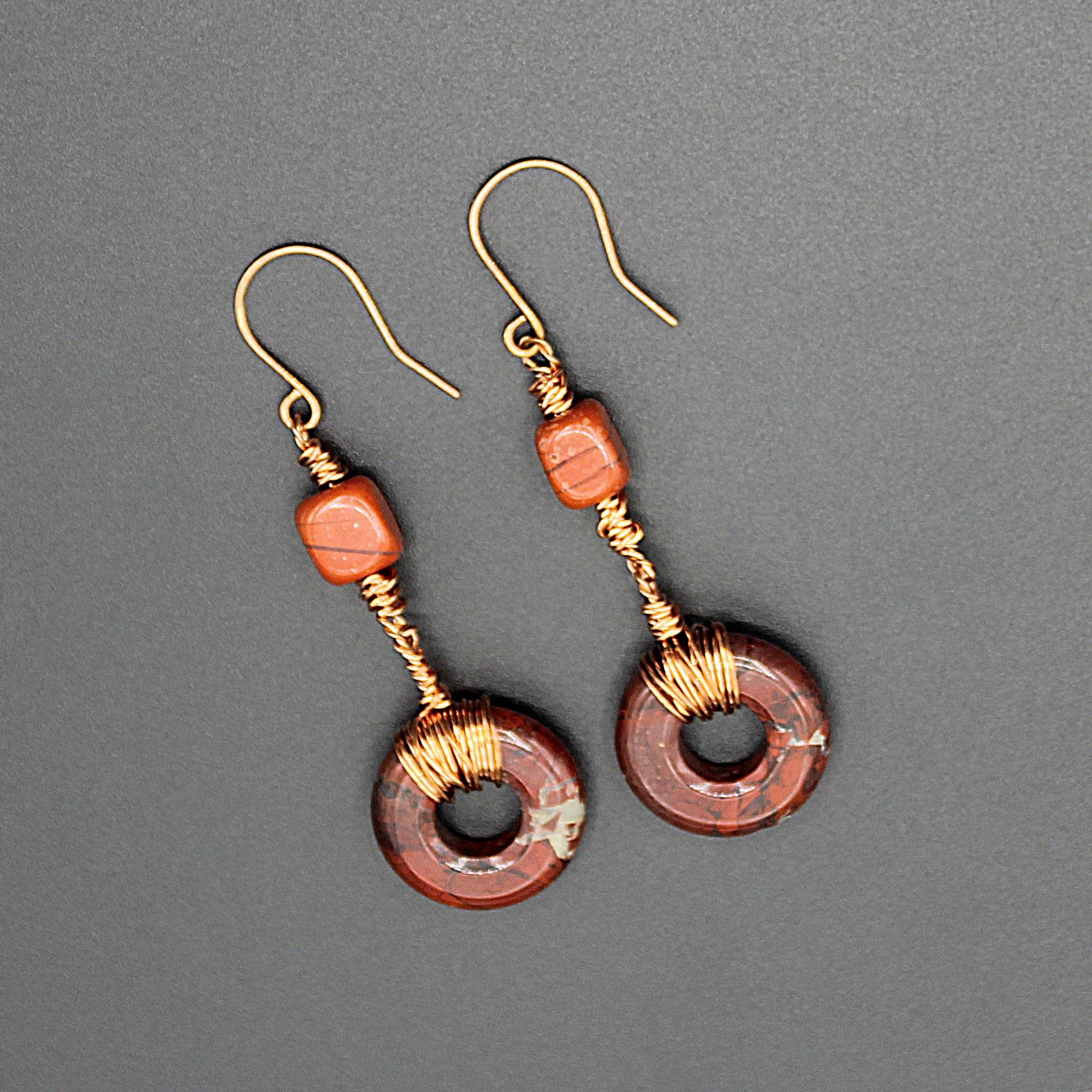 Pair of earrings with donut-shaped brownish-red stones that have striations, wrapped with copper  wire on a gray background