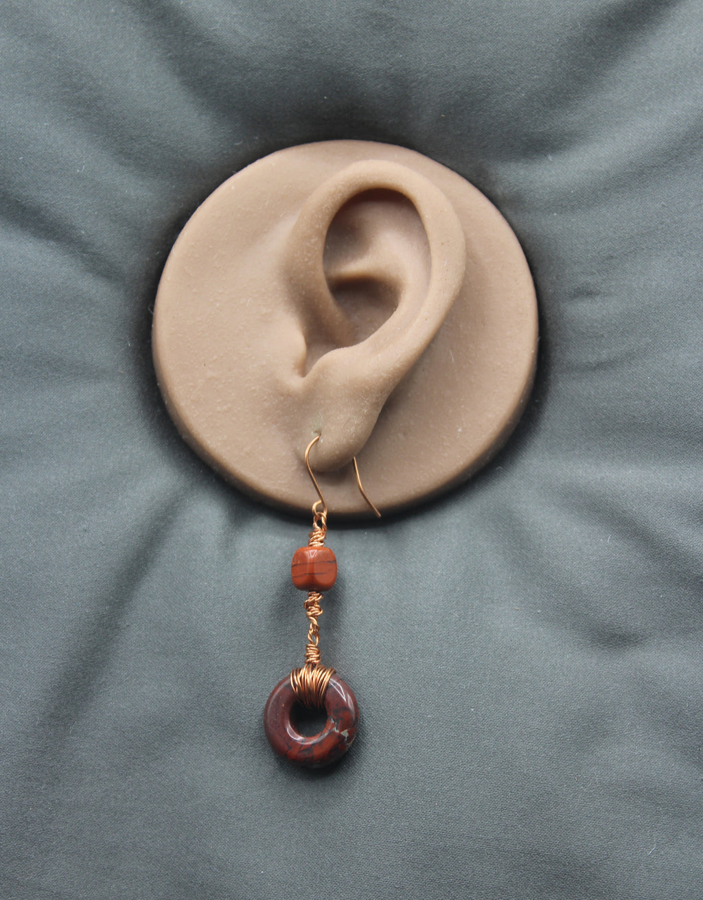 An earring with a donut-shaped brownish-red stone wrapped with copper wire on a clay model of an ear against a gray background