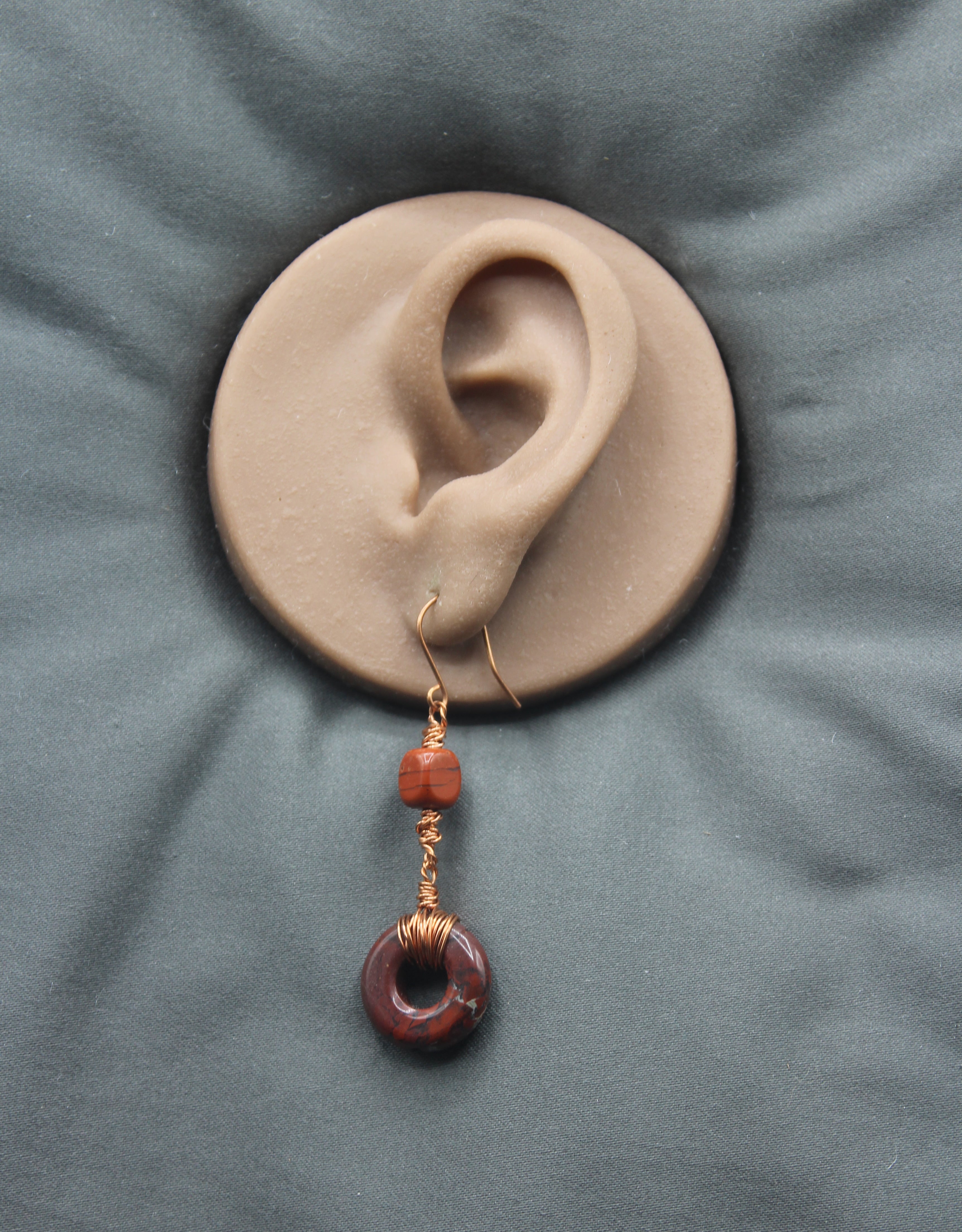 An earring with a donut-shaped brownish-red stone wrapped with copper wire on a clay model of an ear against a gray background