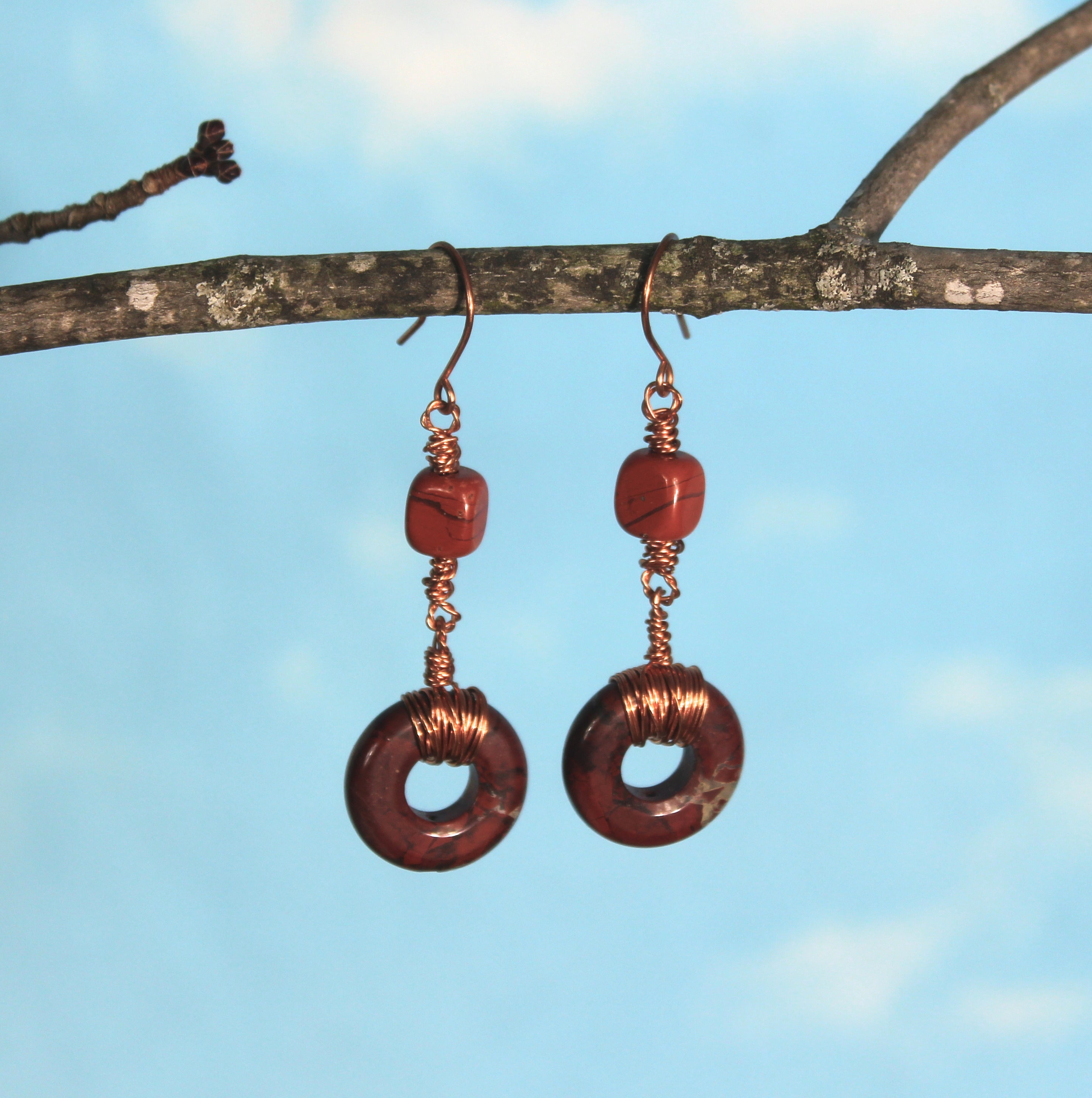 Copper and brownish-red beaded earrings hanging from a branch against a blue sky.