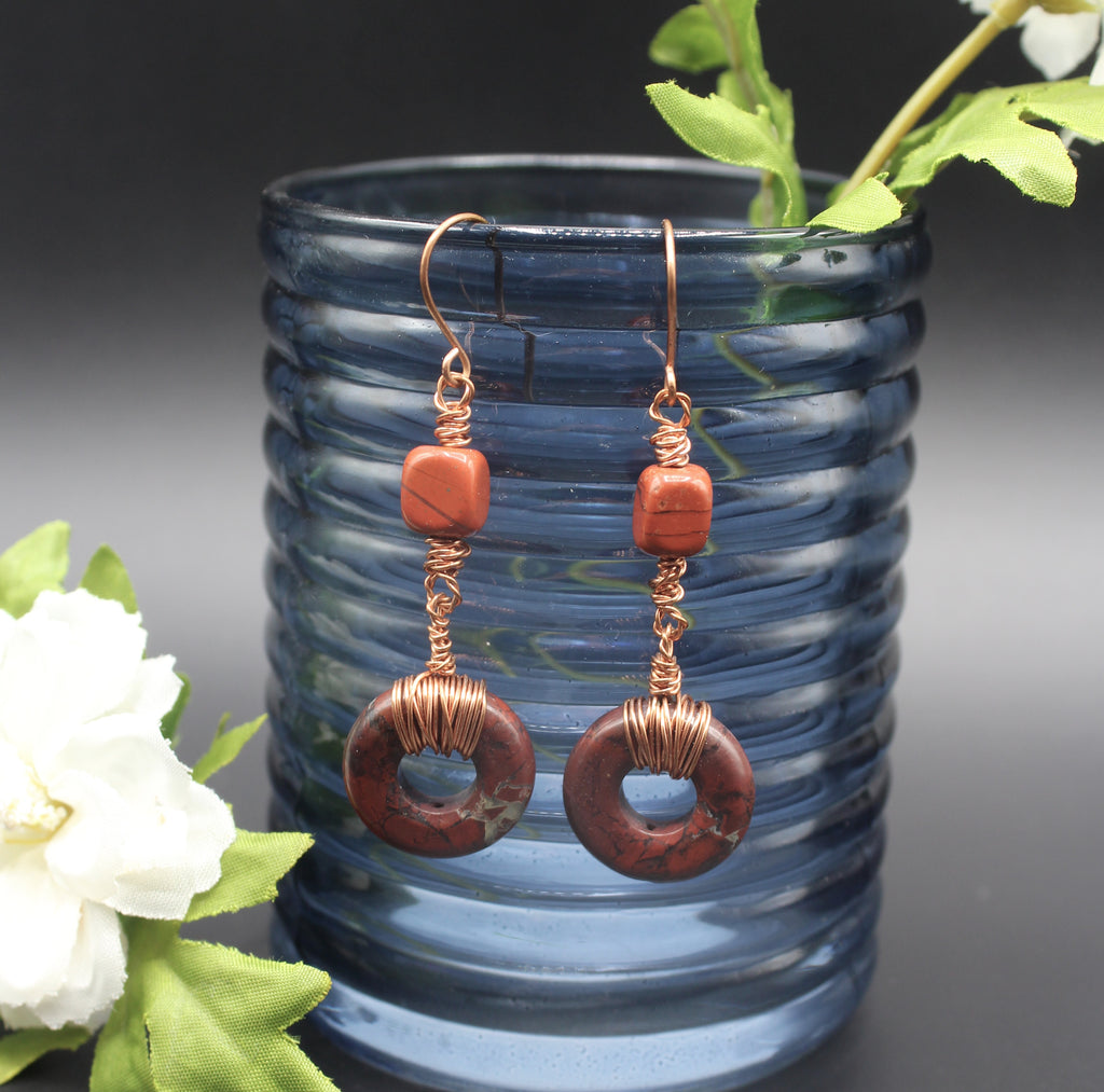 Pair of earrings with donut-shaped brownish-red stones that have striations, wrapped with copper wire, hanging from a blue glass vase.