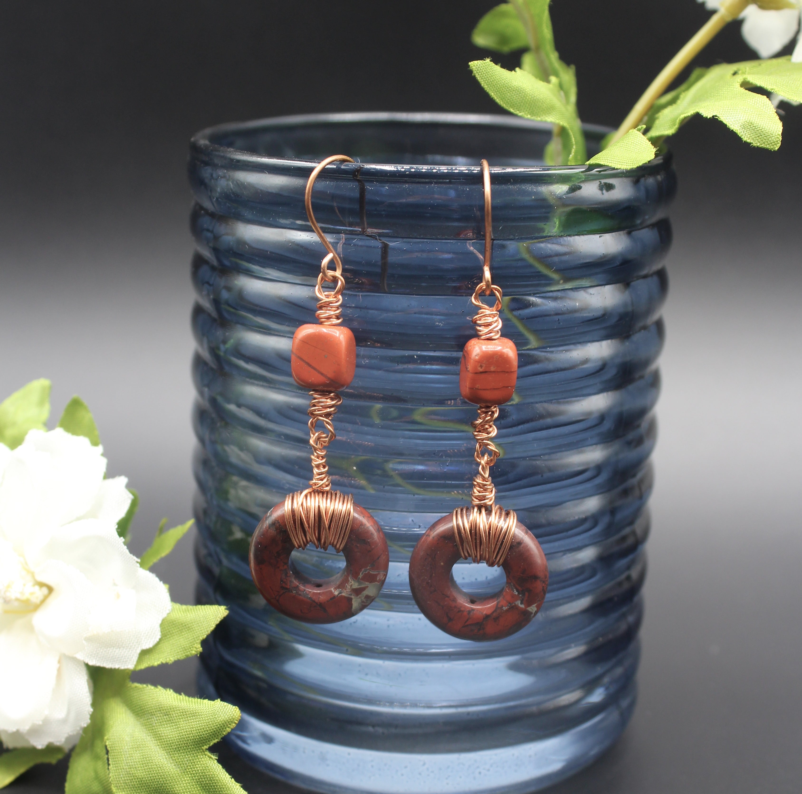Pair of earrings with donut-shaped brownish-red stones that have striations, wrapped with copper wire, hanging from a blue glass vase.