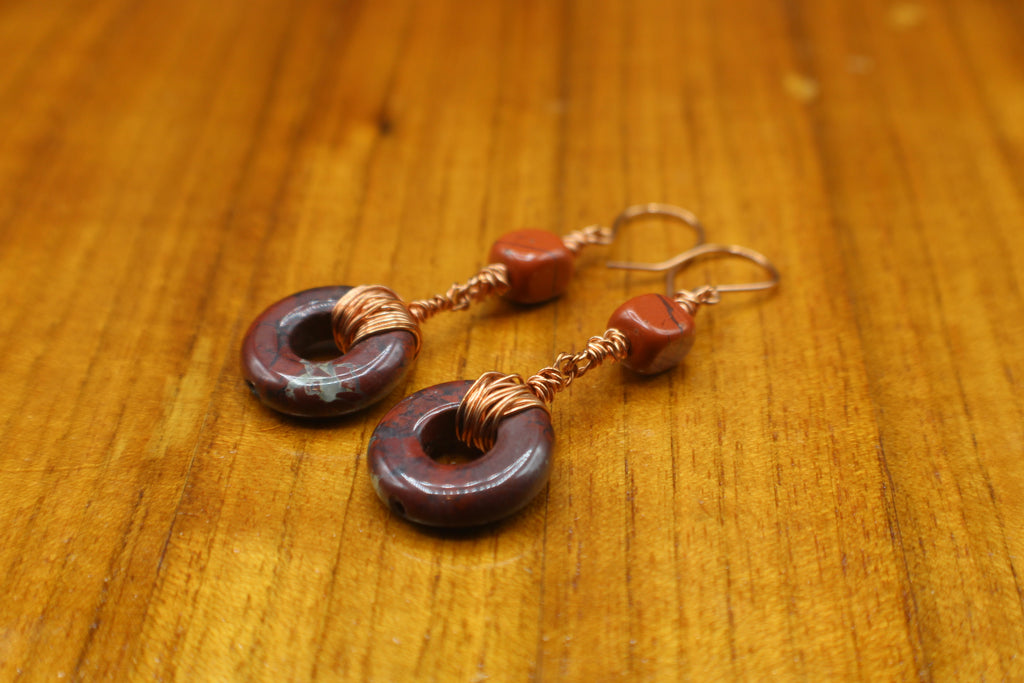 Pair of earrings with donut-shaped brownish-red stones that have striations, wrapped with copper wire on a gray background on a wooden surface