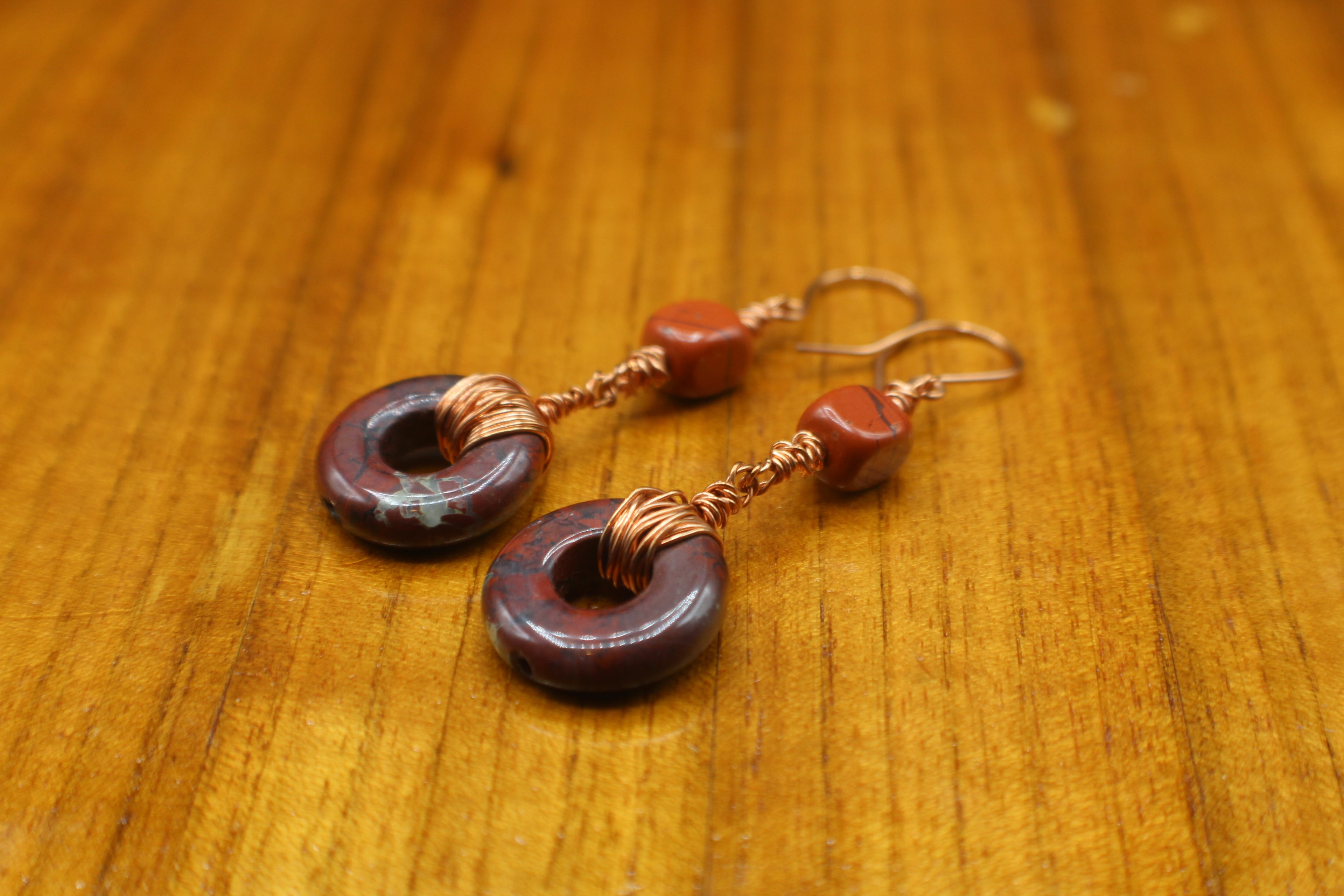 Pair of earrings with donut-shaped brownish-red stones that have striations, wrapped with copper wire on a gray background on a wooden surface