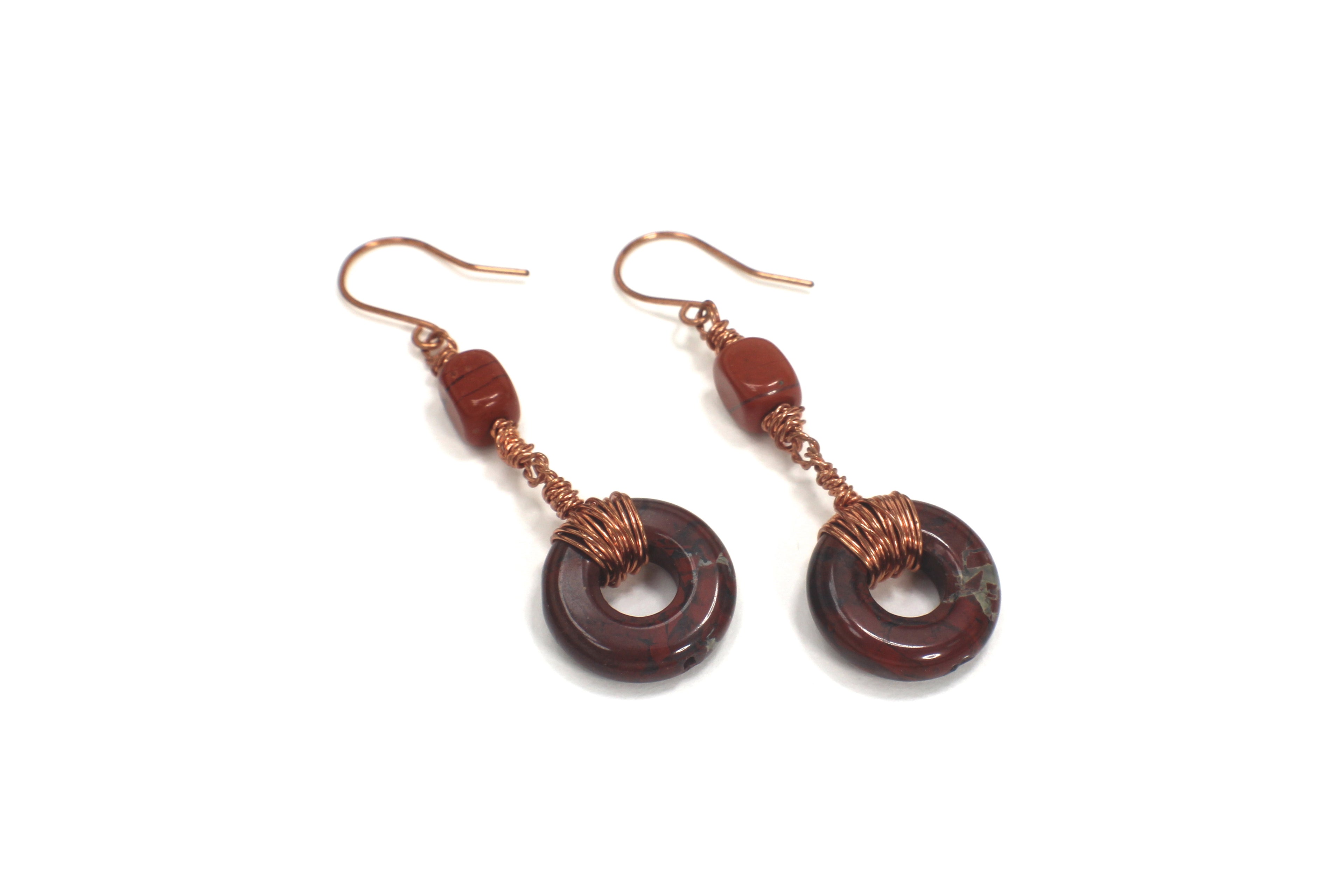 Pair of earrings with donut-shaped brownish-red stones that have striations, wrapped with copper wire on a white background