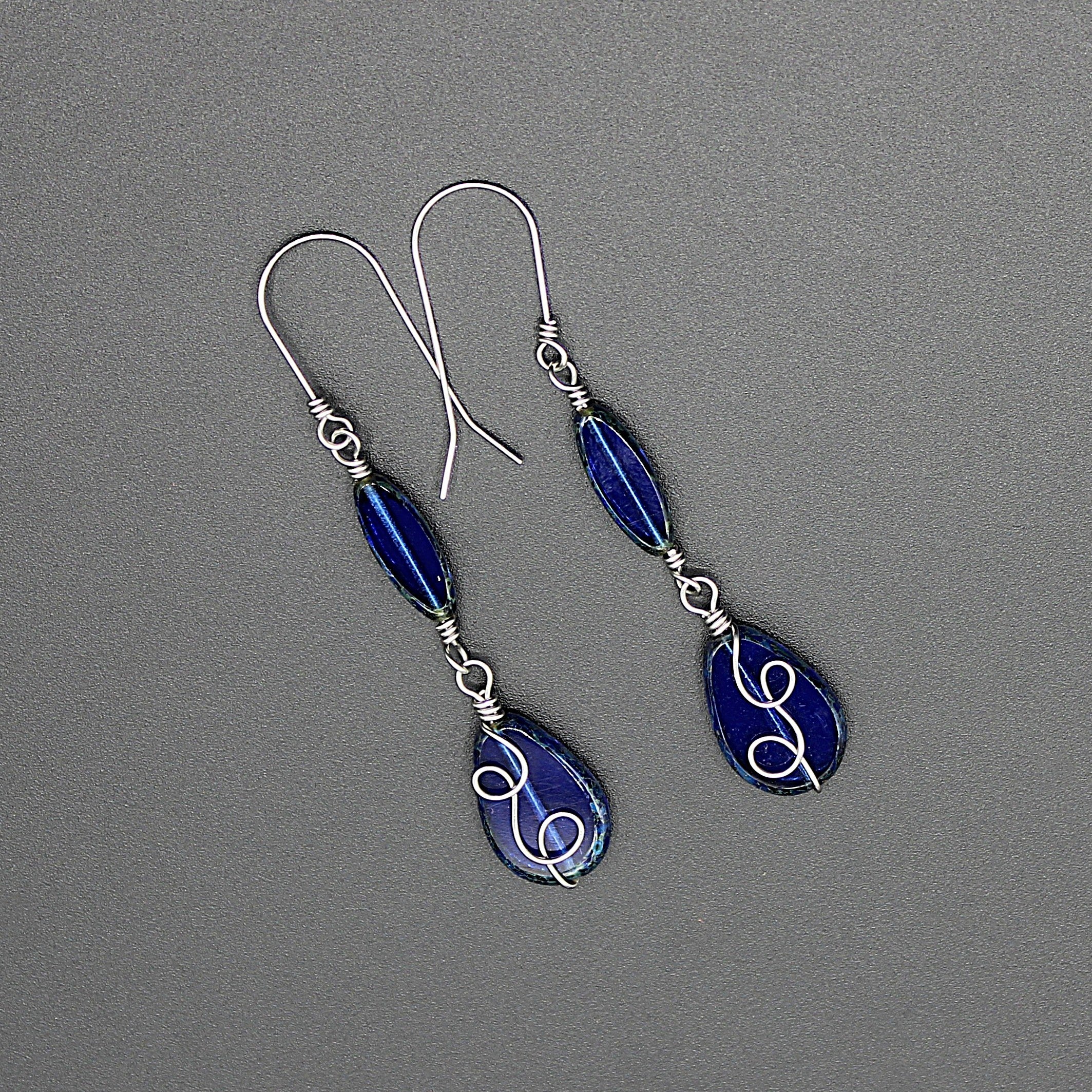 Pair of sapphire blue teardrop earrings with stainless steel wire details on a gray background