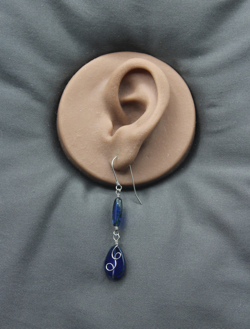 Sapphire blue teardrop earring with stainless steel wire details on a clay model of an ear