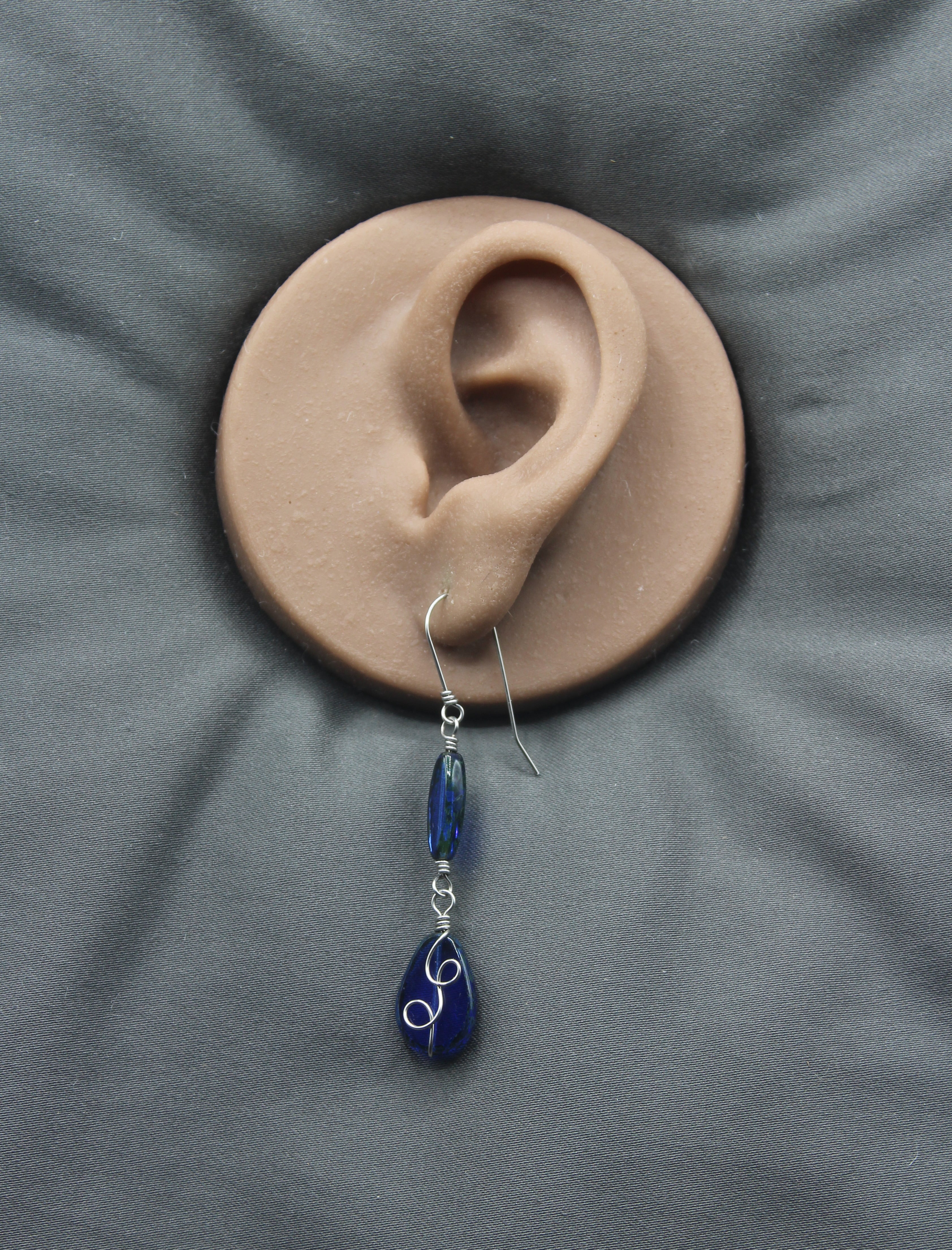 Sapphire blue teardrop earring with stainless steel wire details on a clay model of an ear