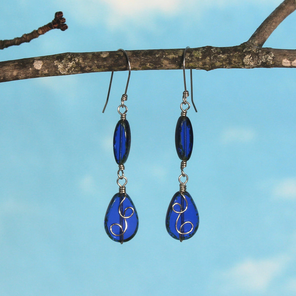 Sapphire blue glass drop earrings with stainless steel wire details hanging from a branch against a blue sky.