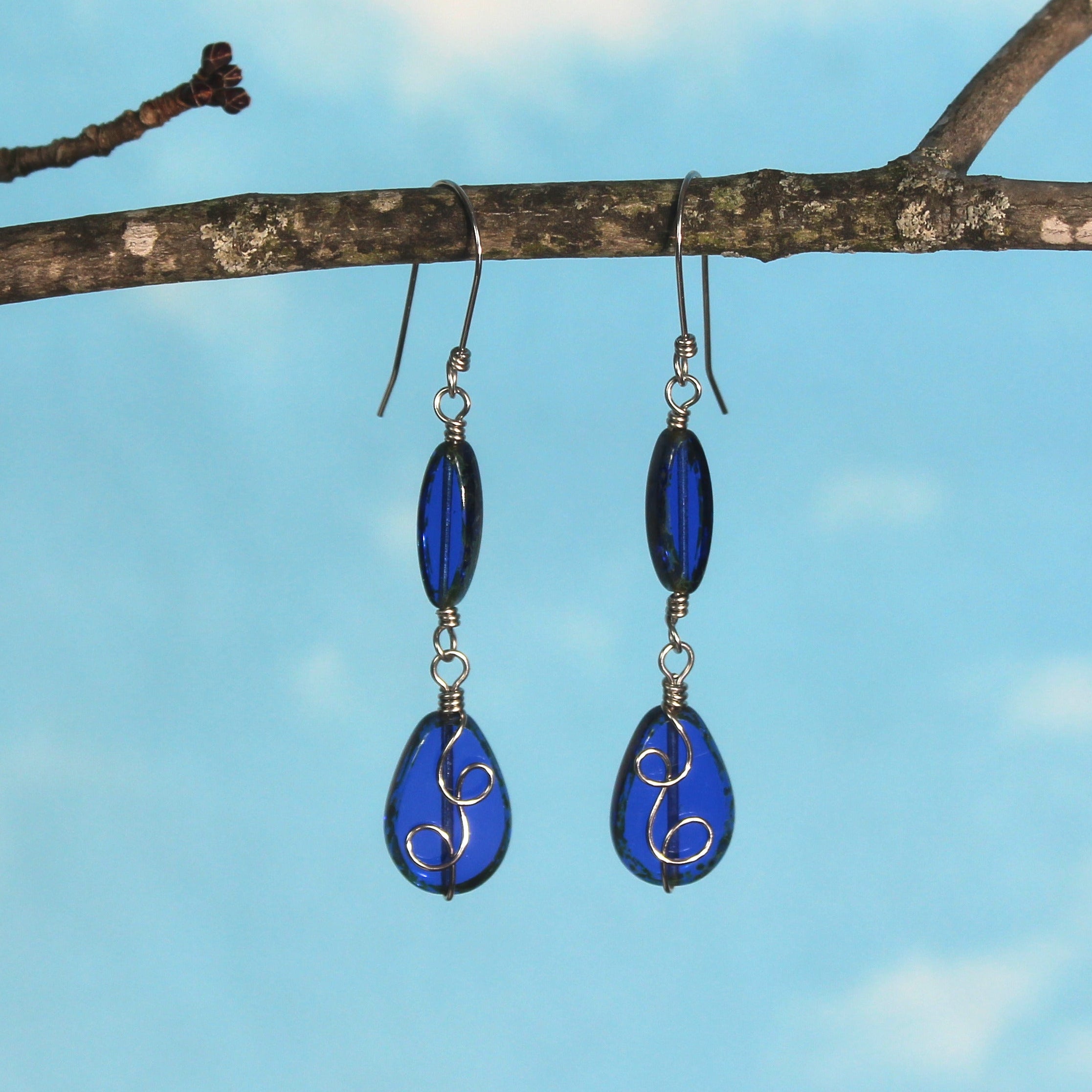 Sapphire blue glass drop earrings with stainless steel wire details hanging from a branch against a blue sky.