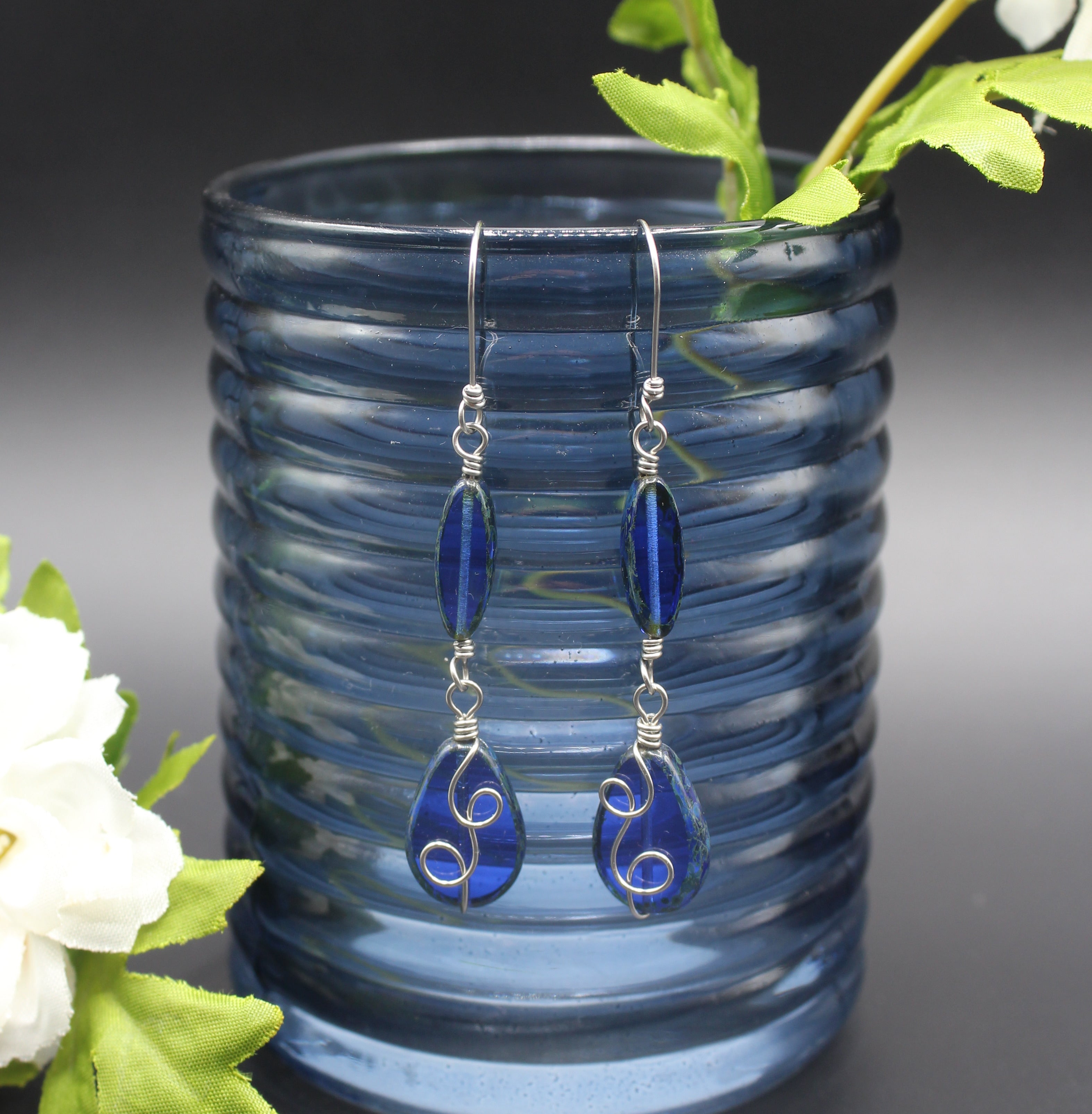Sapphire blue glass earrings with stainless steel wire details hanging from a blue glass vase.