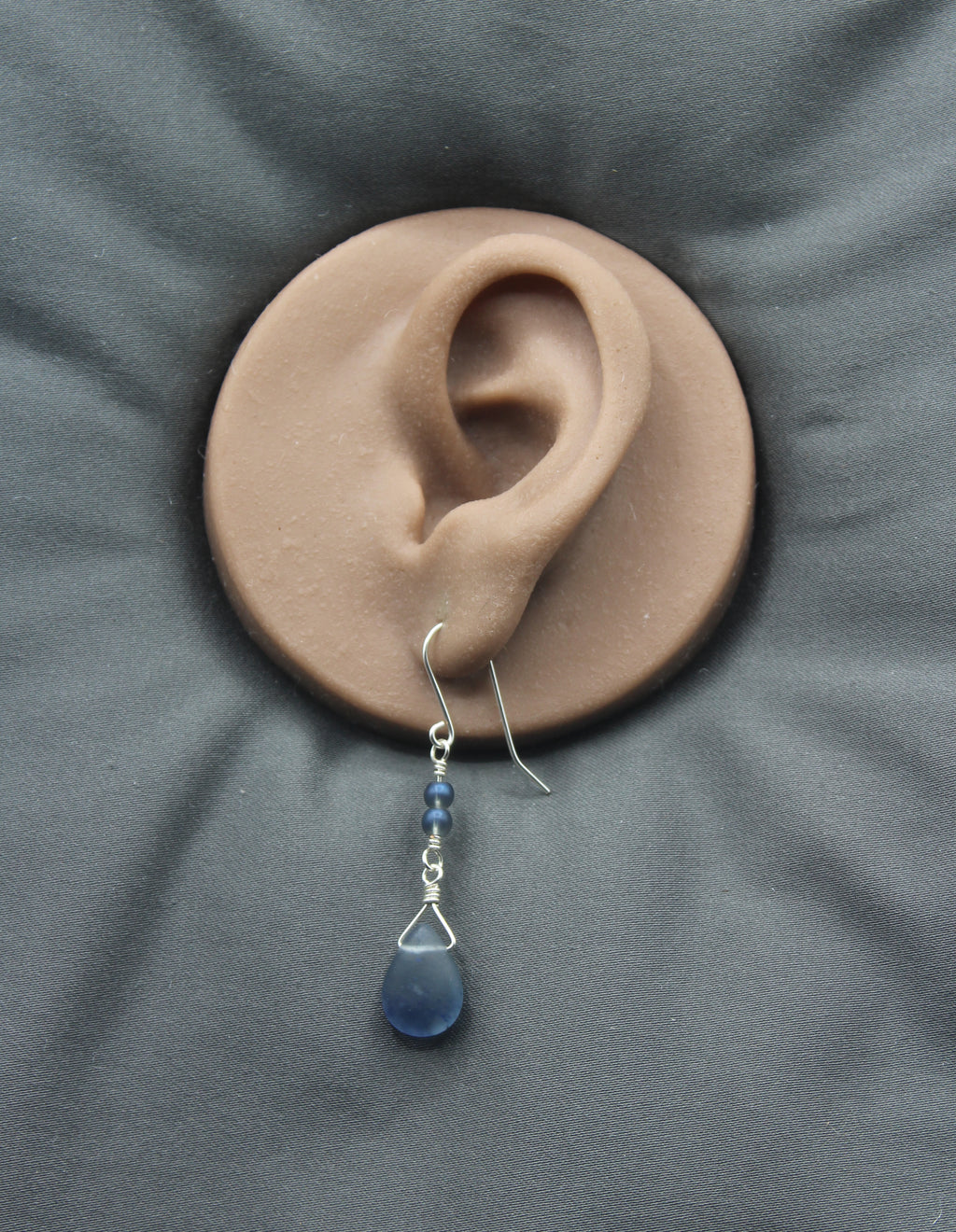 Blue teardrop earring on a clay model of an ear with a gray background