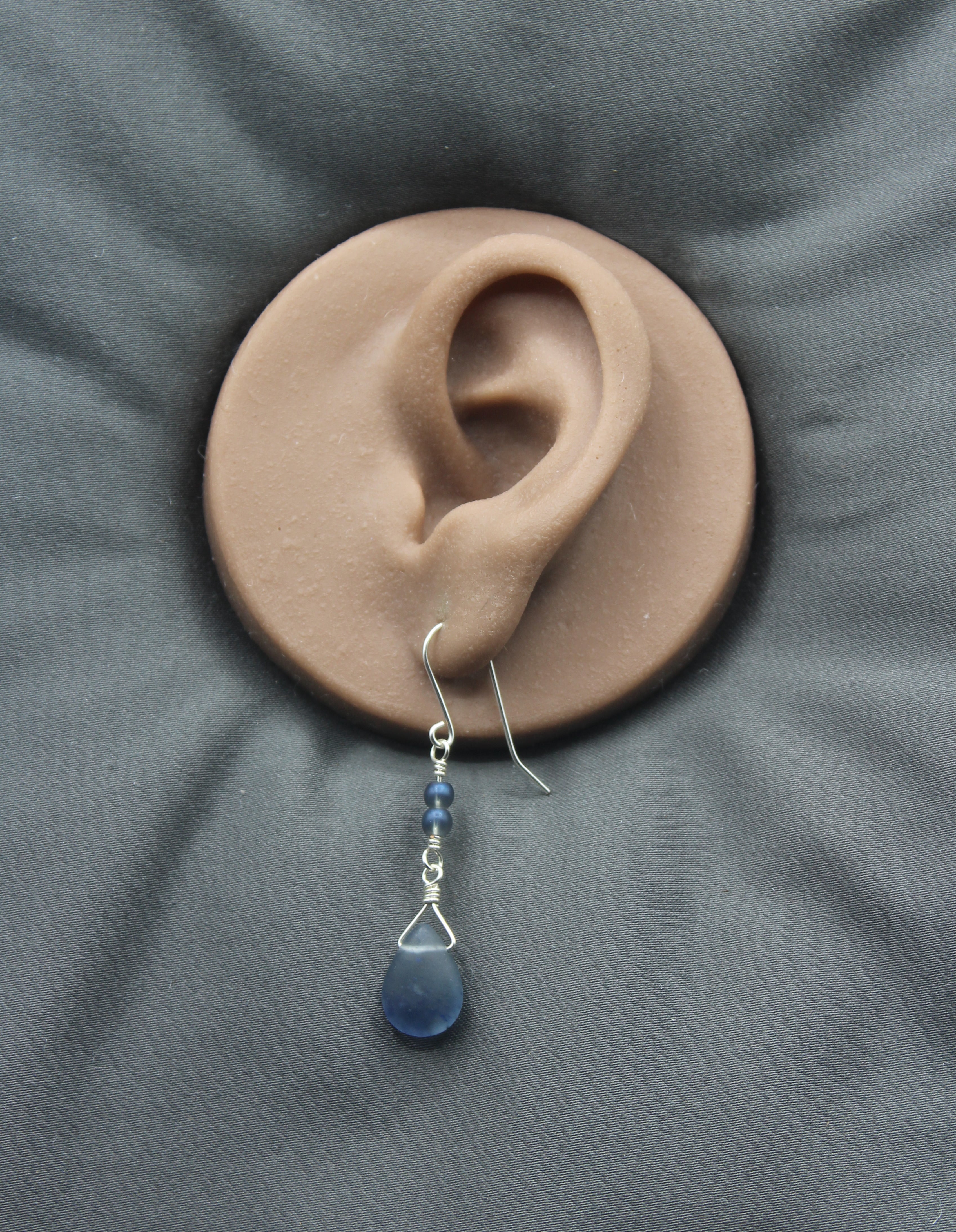Blue teardrop earring on a clay model of an ear with a gray background