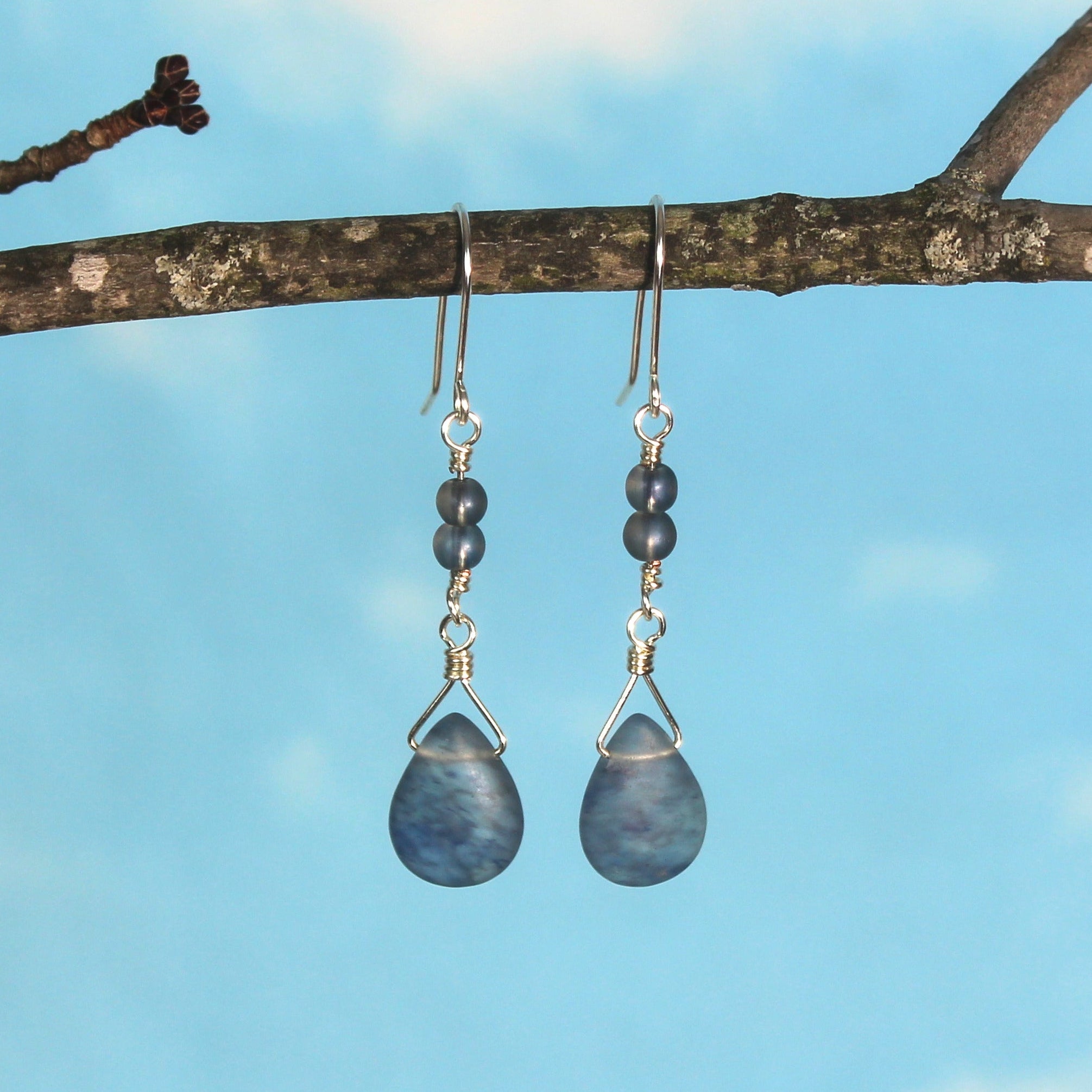 Blue teardrop earrings hanging from a branch with a blue sky background